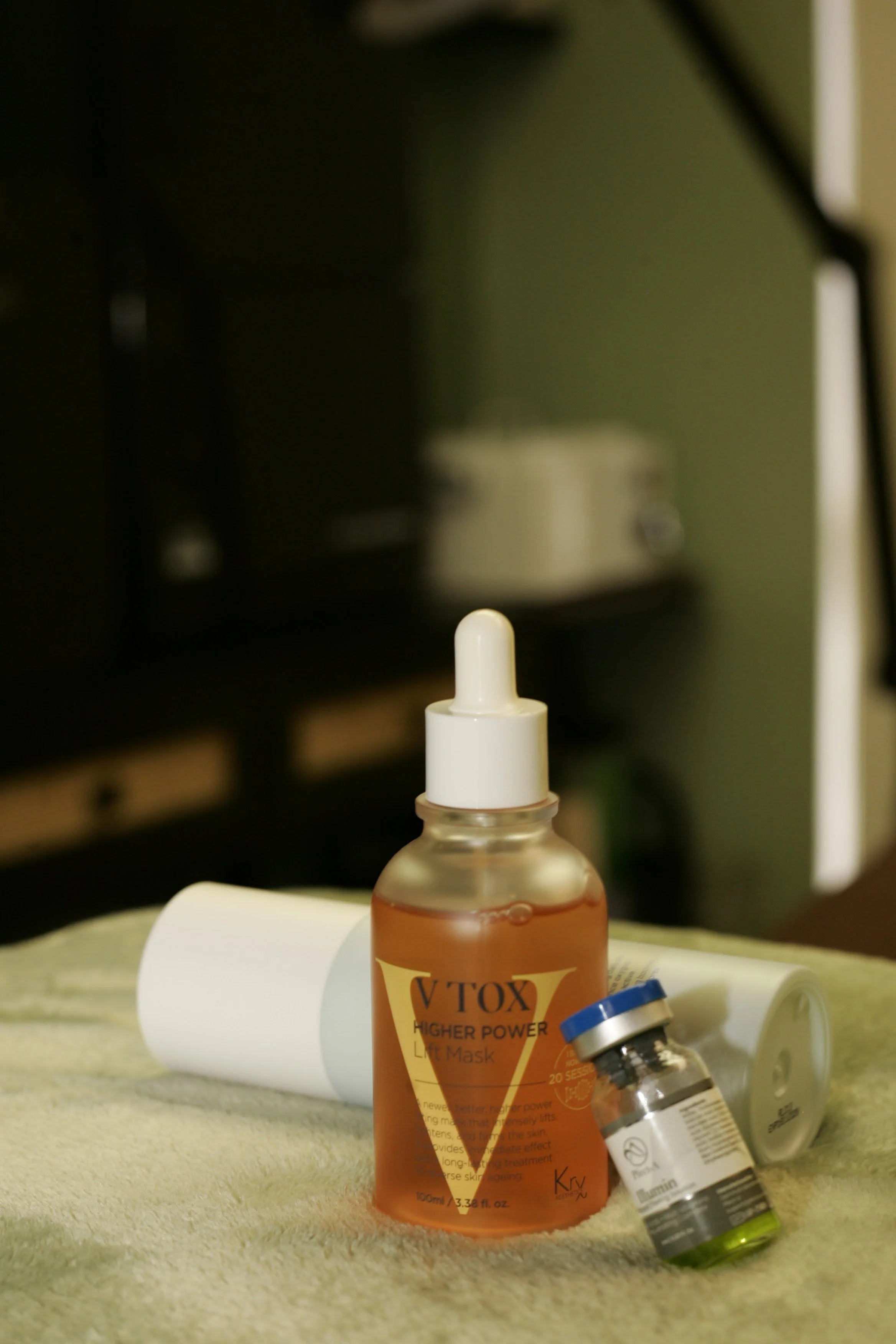 Assorted bottles and tubes of skincare and medication products on a beige surface, with a blurry background including a green wall and a black object.