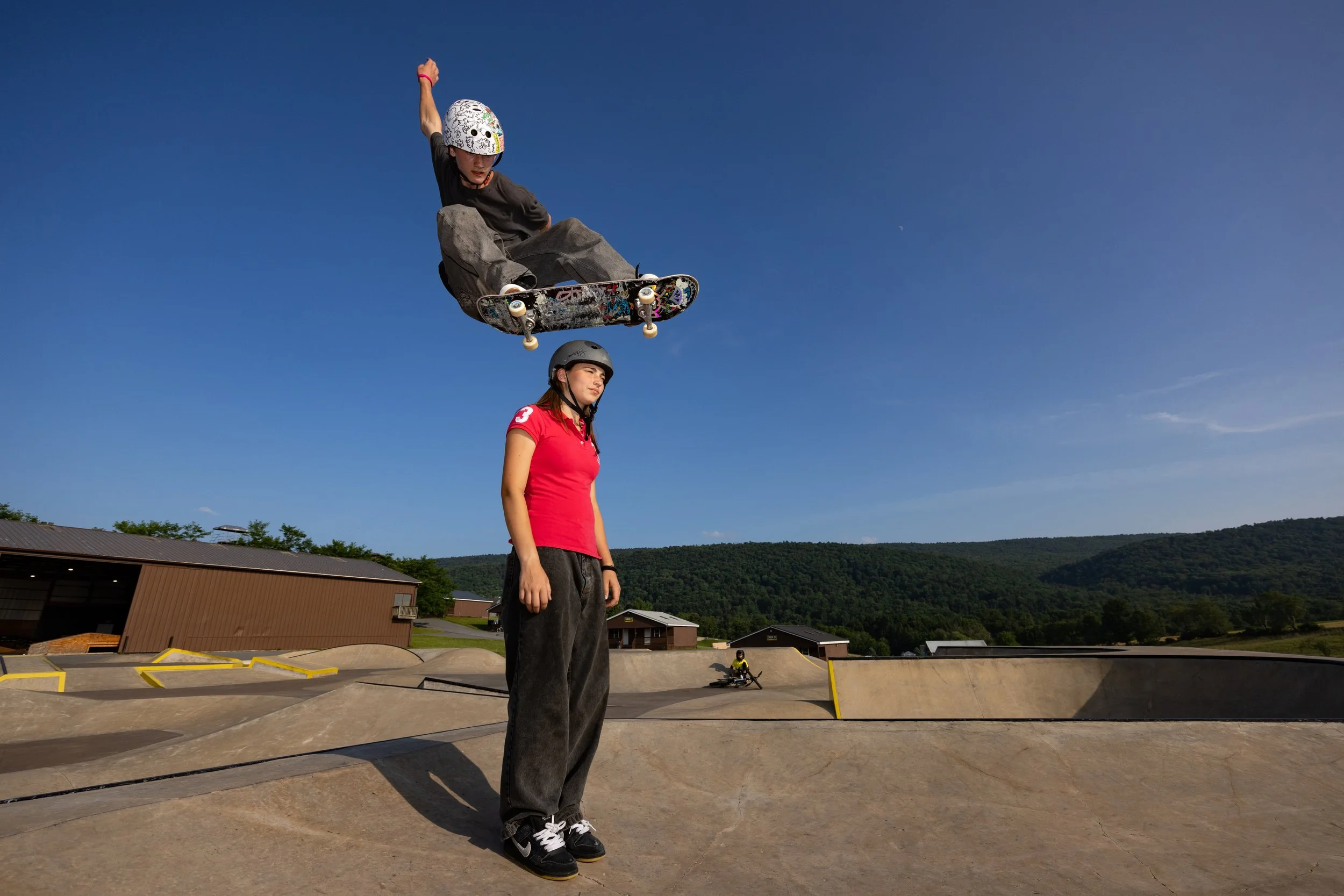 A person at a skate park performing a jump on a skateboard, wearing a helmet and casual clothes, with a clear blue sky overhead and other skateboarders in the background.
