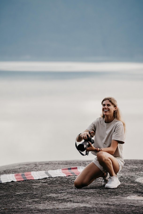 A young woman with blonde hair, wearing a gray t-shirt and shorts, kneeling on a rock with a camera in hand, smiling near a body of water with a blanket on the ground.