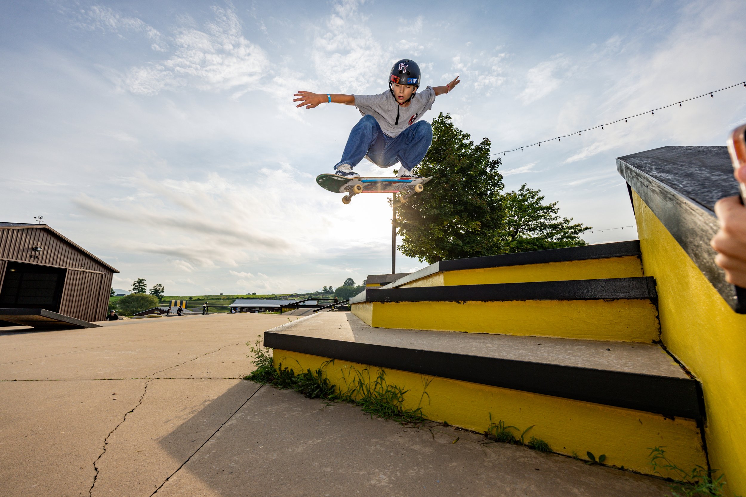A young boy skateboard jumps off a yellow ledge at a skate park during daytime, wearing a helmet and casual clothing, with a tree and cloudy sky in the background.
