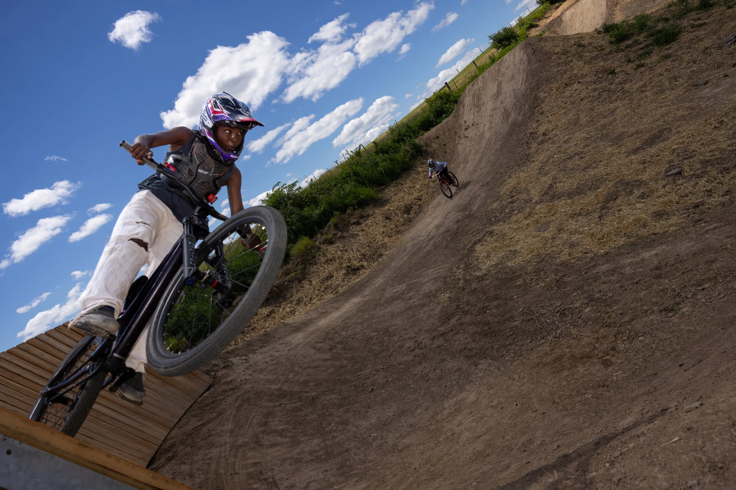 Two young children riding mountain bikes downhill on a dirt trail under a blue sky with white clouds, surrounded by greenery.