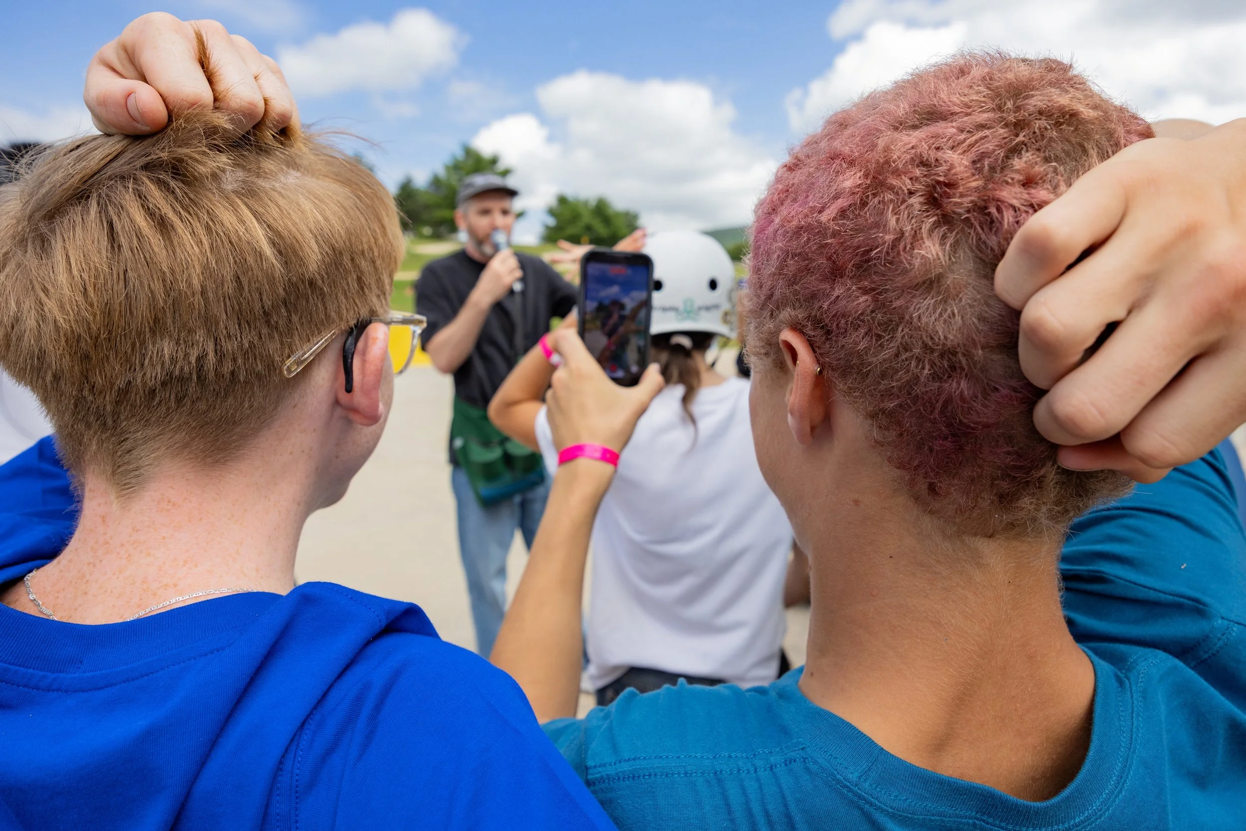 Close-up of two people with dyed hair, one with blond and the other with pink, at an outdoor event. In the background, a person is speaking into a microphone, and another person is taking a photo with a smartphone. The sky is partly cloudy, and there are people and trees in the distance.