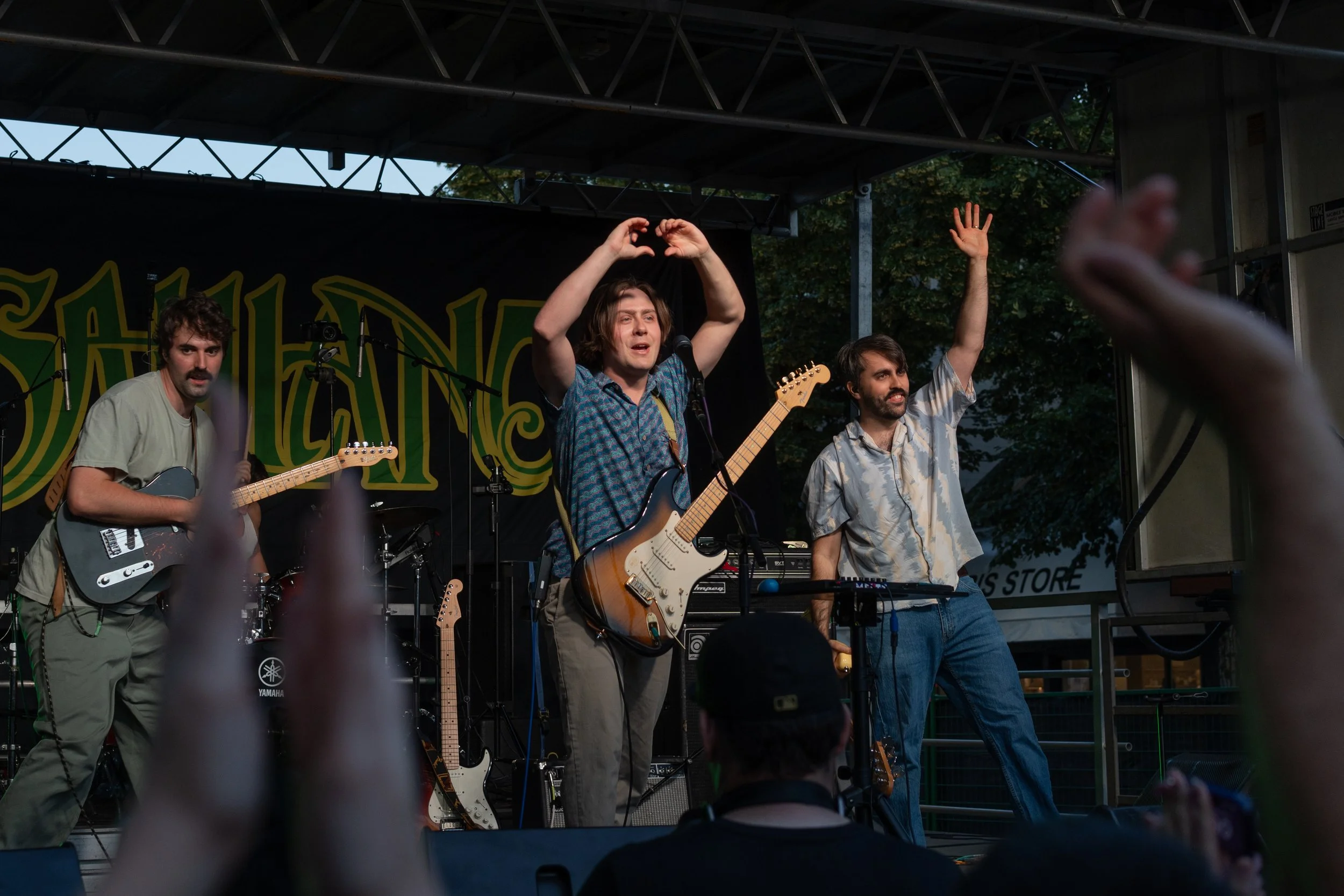 Four musicians performing on an outdoor stage. One is playing a guitar, another is singing with a microphone, and a third is raising his hand. The audience is visible with raised hands in the foreground.