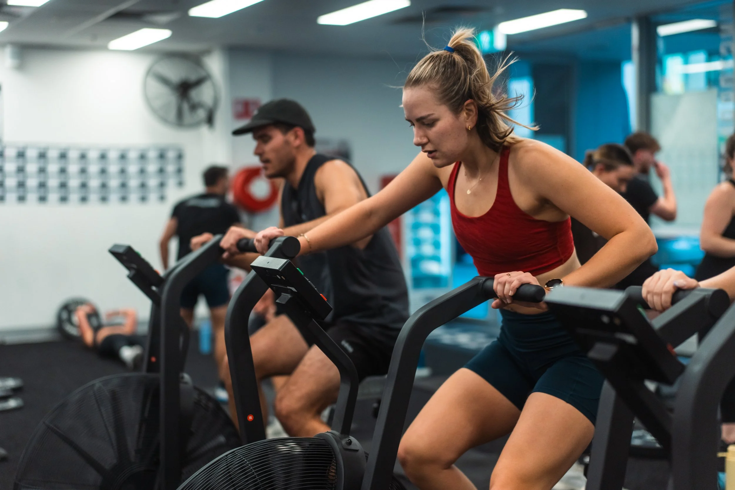 People exercising on stationary bikes at a gym with a large fan and blue gym walls.