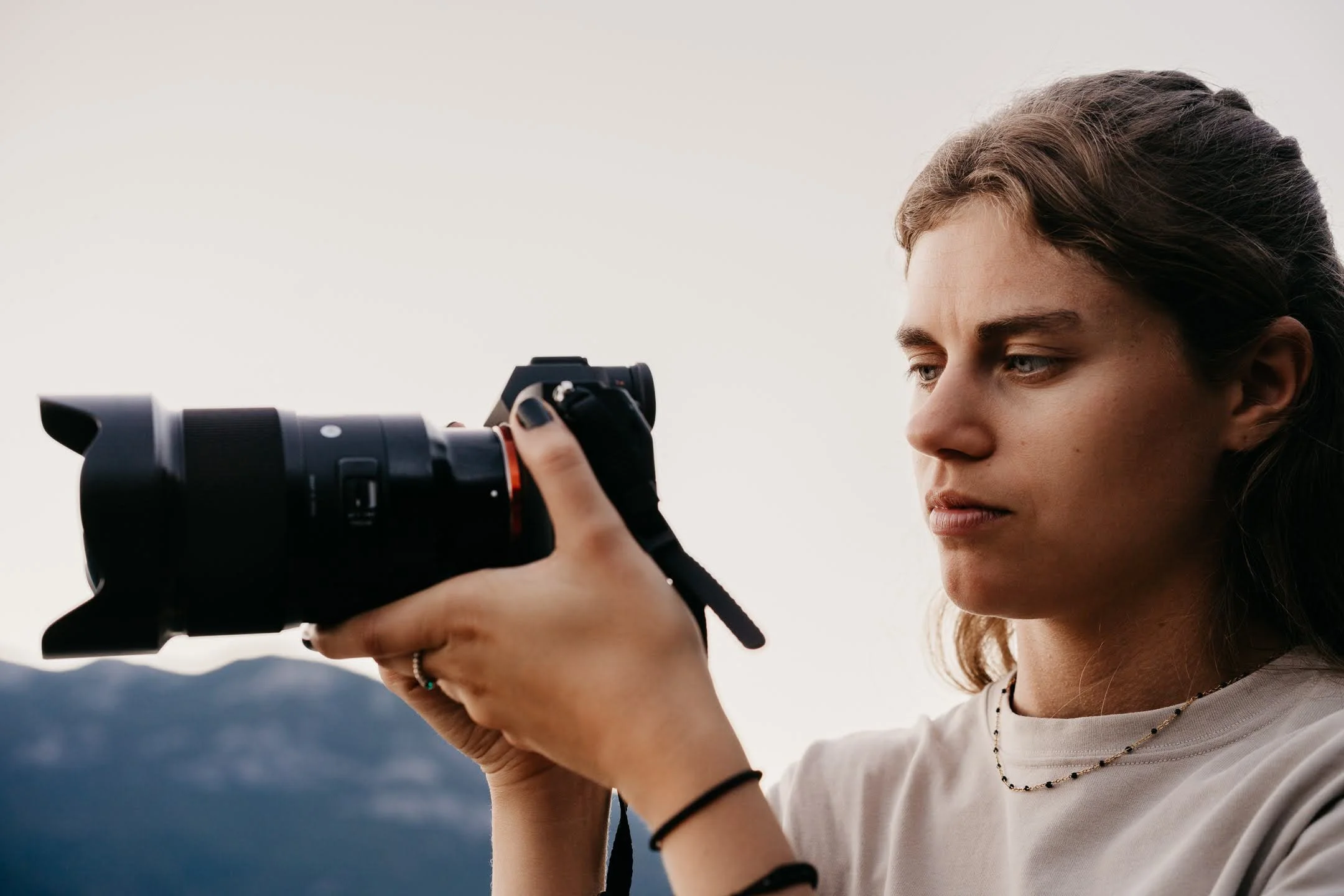A young woman with brown hair tied back is holding a camera up to take a photo, with a blurred natural landscape in the background.