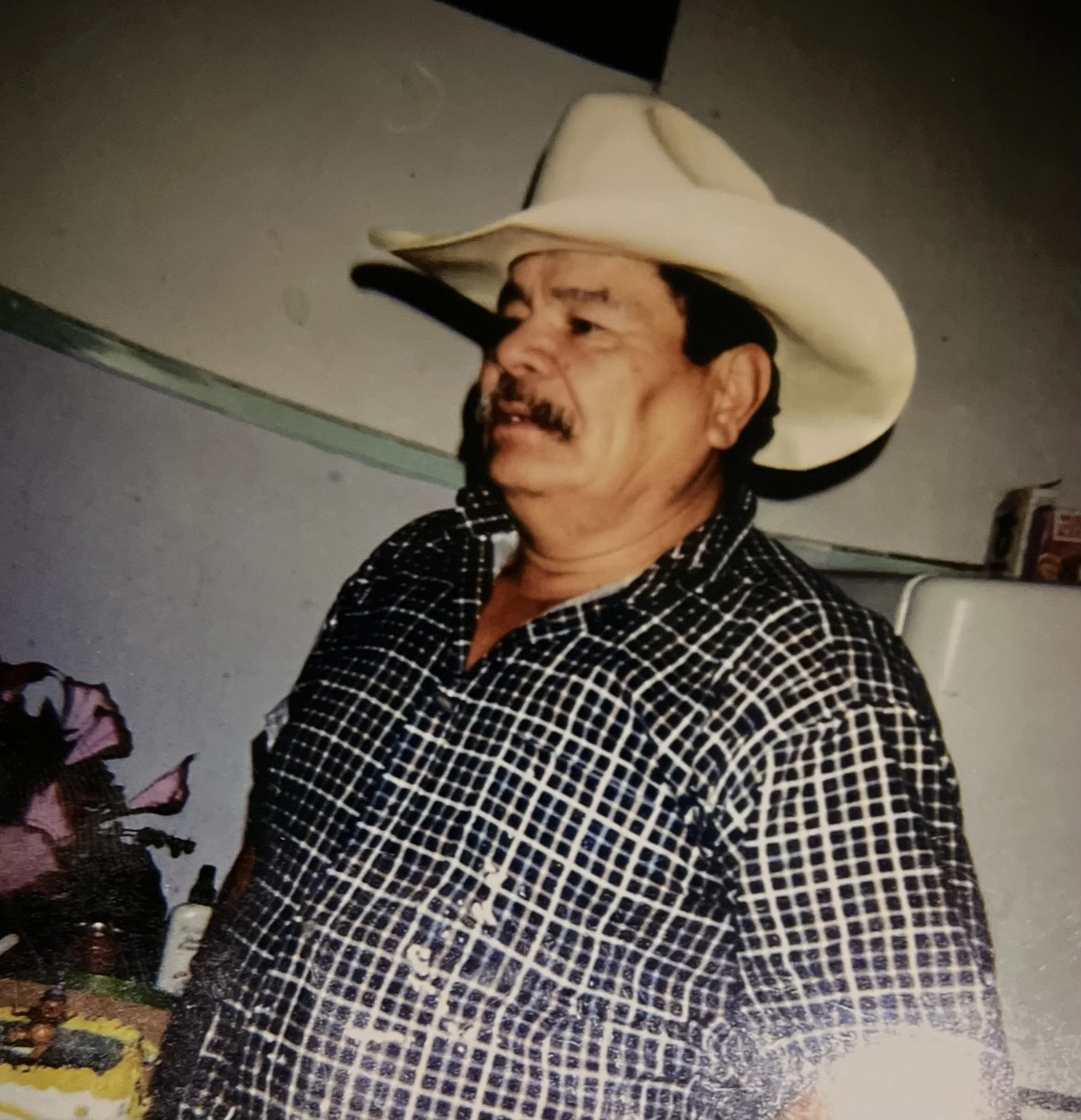 A man wearing a black and white checkered shirt and a big white cowboy hat, with a dark mustache, standing against a light-colored wall.