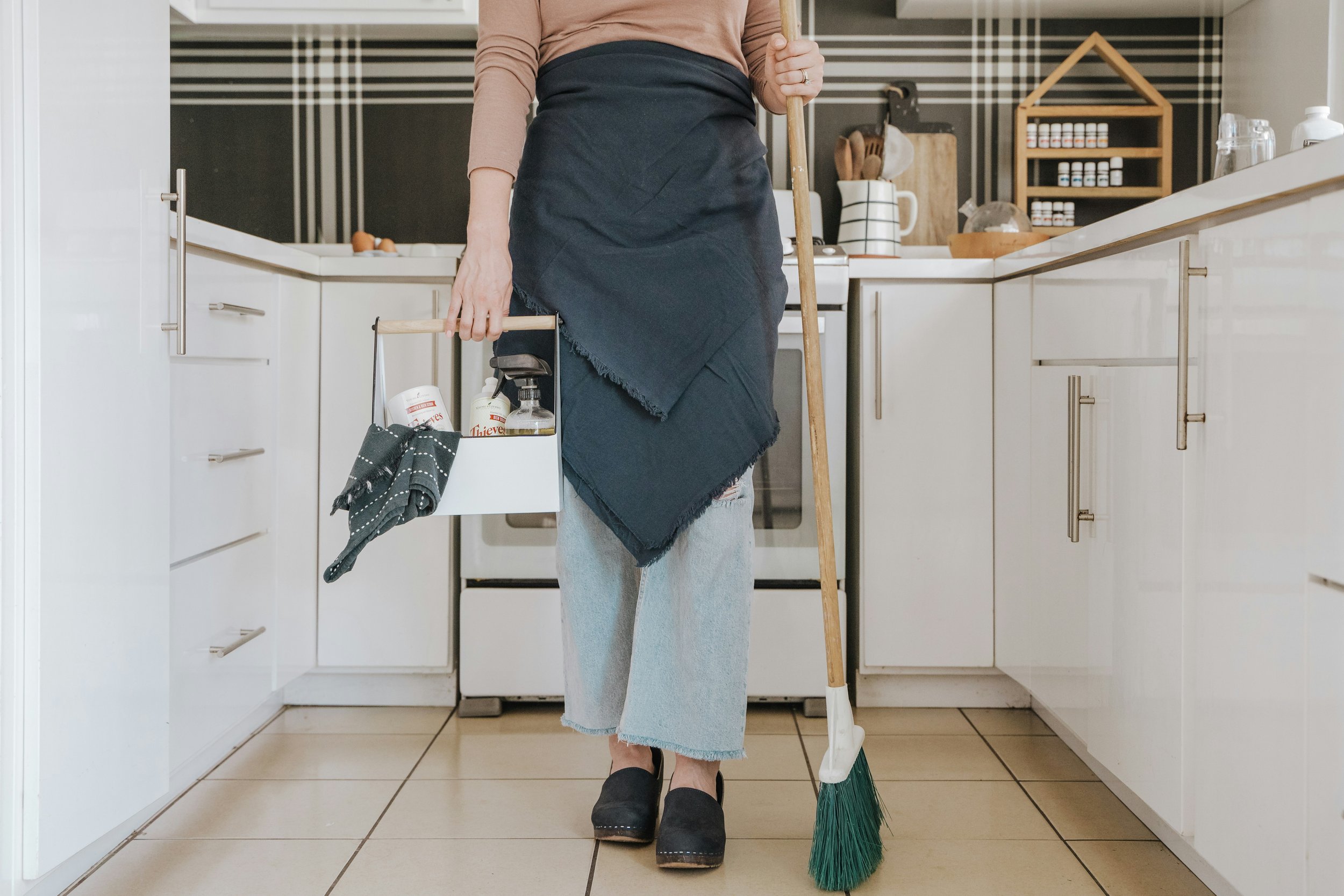 A person standing in a kitchen holding a mop and a bucket, wearing a blue apron and slippers. The kitchen has white cabinets, tile flooring, and various kitchen items on the counter and shelves.