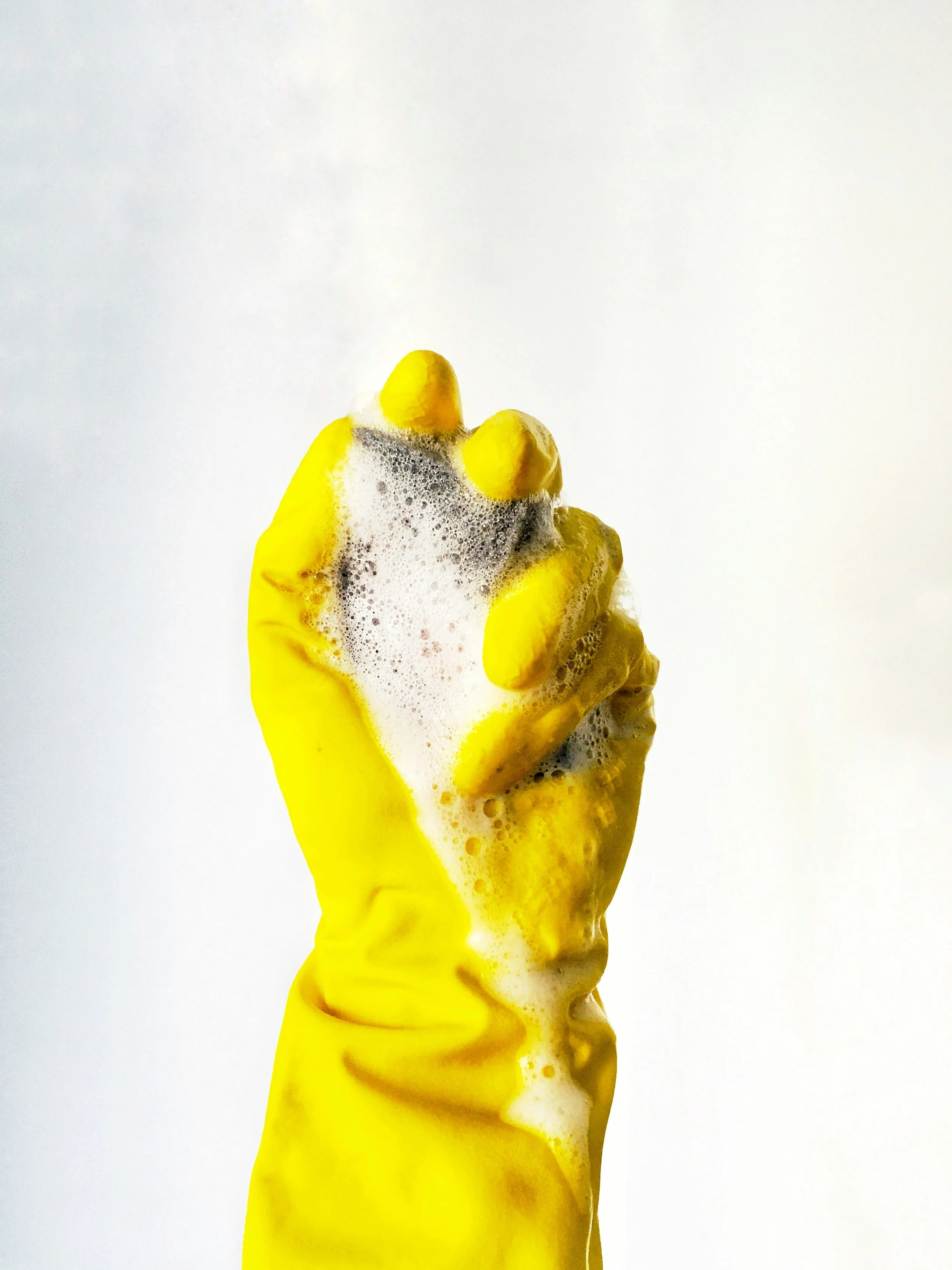 Yellow rubber glove holding a soapy, sudsy sponge against a plain white background.