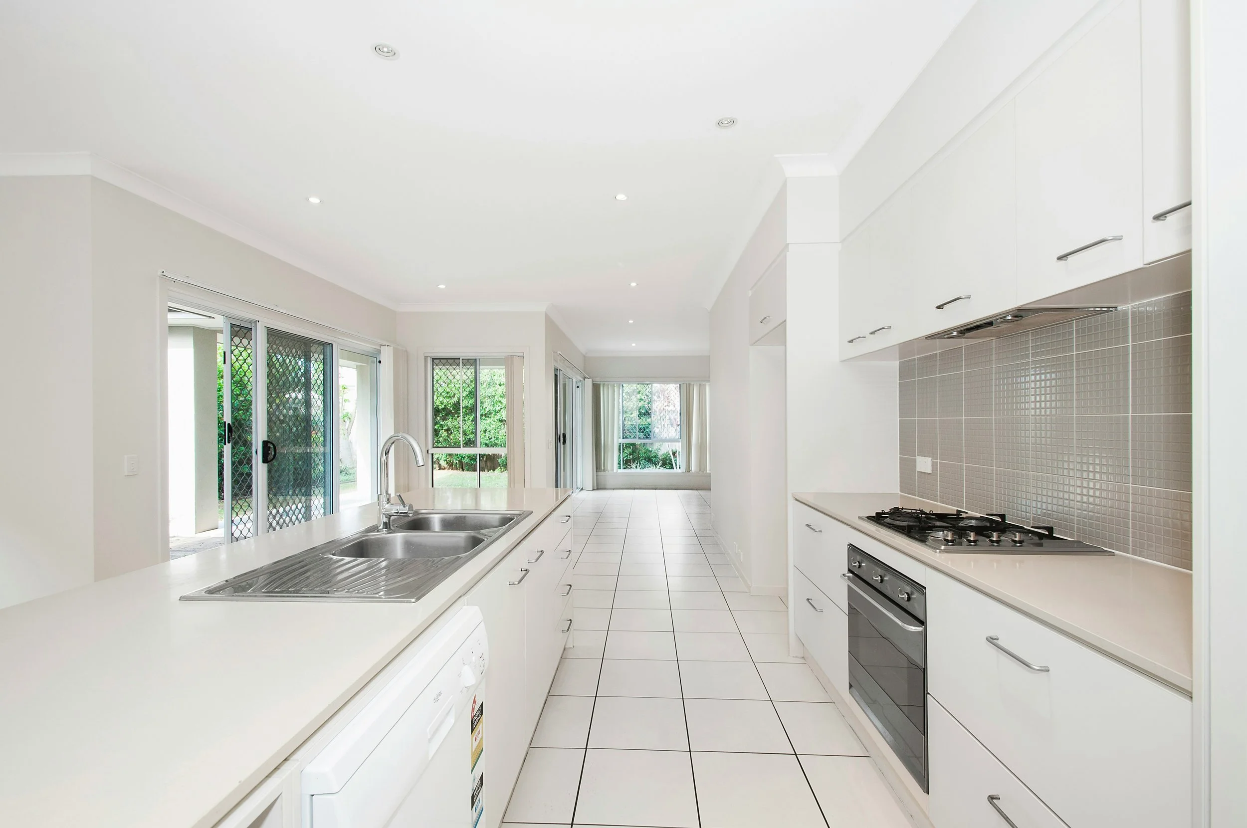 A bright, modern kitchen with white cabinets, a tiled backsplash, a gas stove, a double sink, and large sliding glass doors leading outside to a garden.