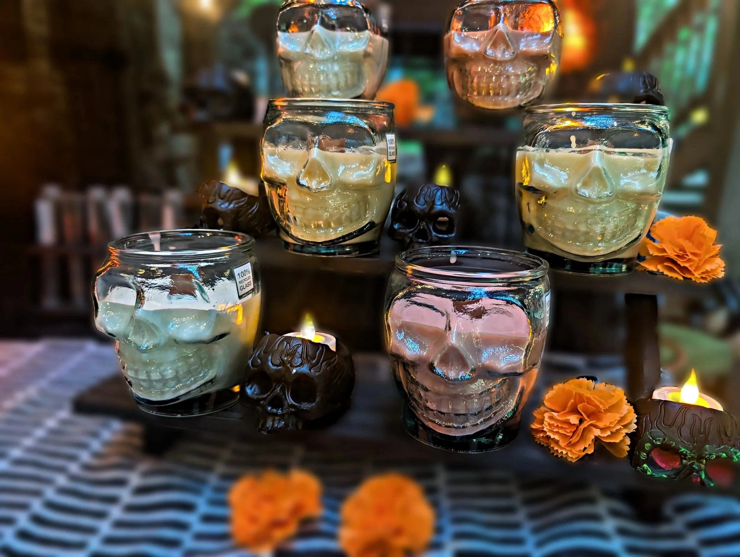 Glass candle holders shaped like skulls with white candles inside, surrounded by small orange marigolds and decorative skulls in the background, illuminated with colorful ambient lighting.