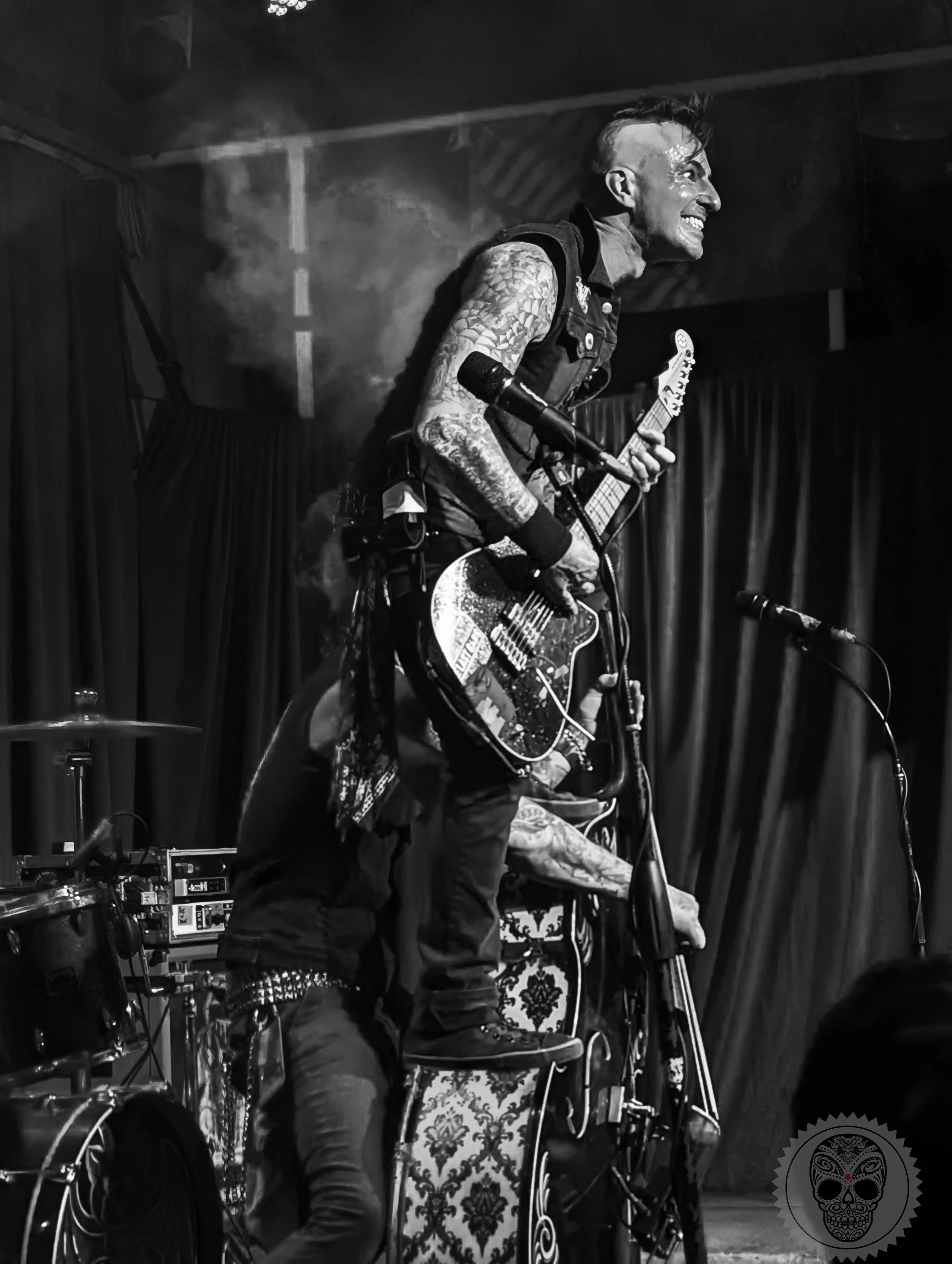 A tattooed man with a mohawk hairstyle playing an electric guitar during a live music performance with a dark curtain backdrop.