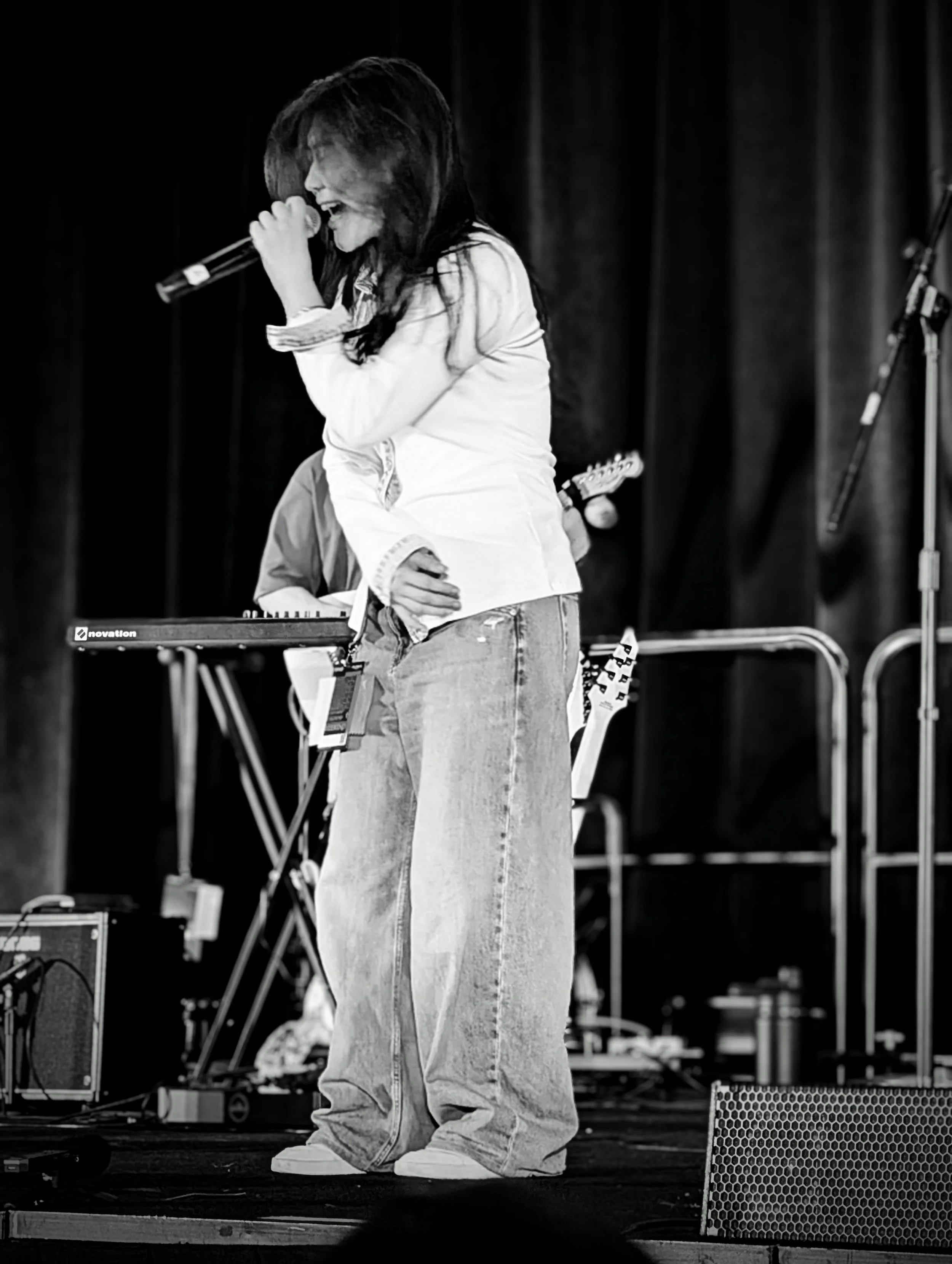 A woman singing into a microphone on stage, with musical equipment and dark curtains in the background.