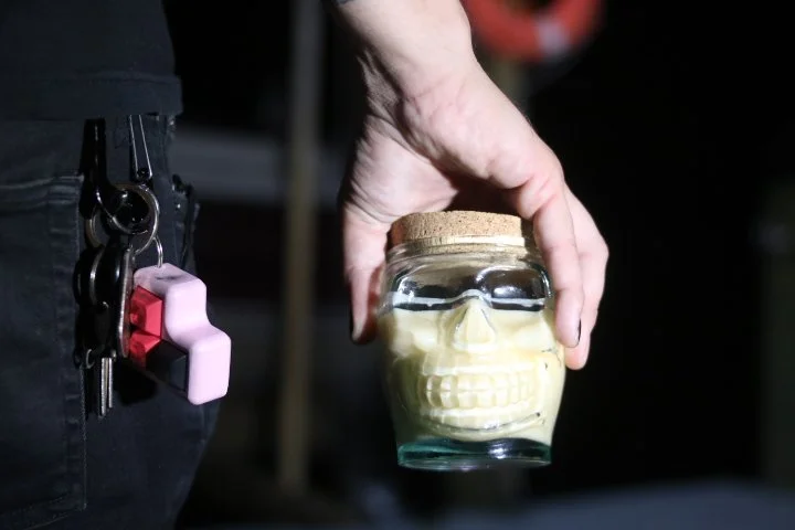 A person's hand holding a glass jar with a cork lid, containing a skull-shaped object, in a dark setting.