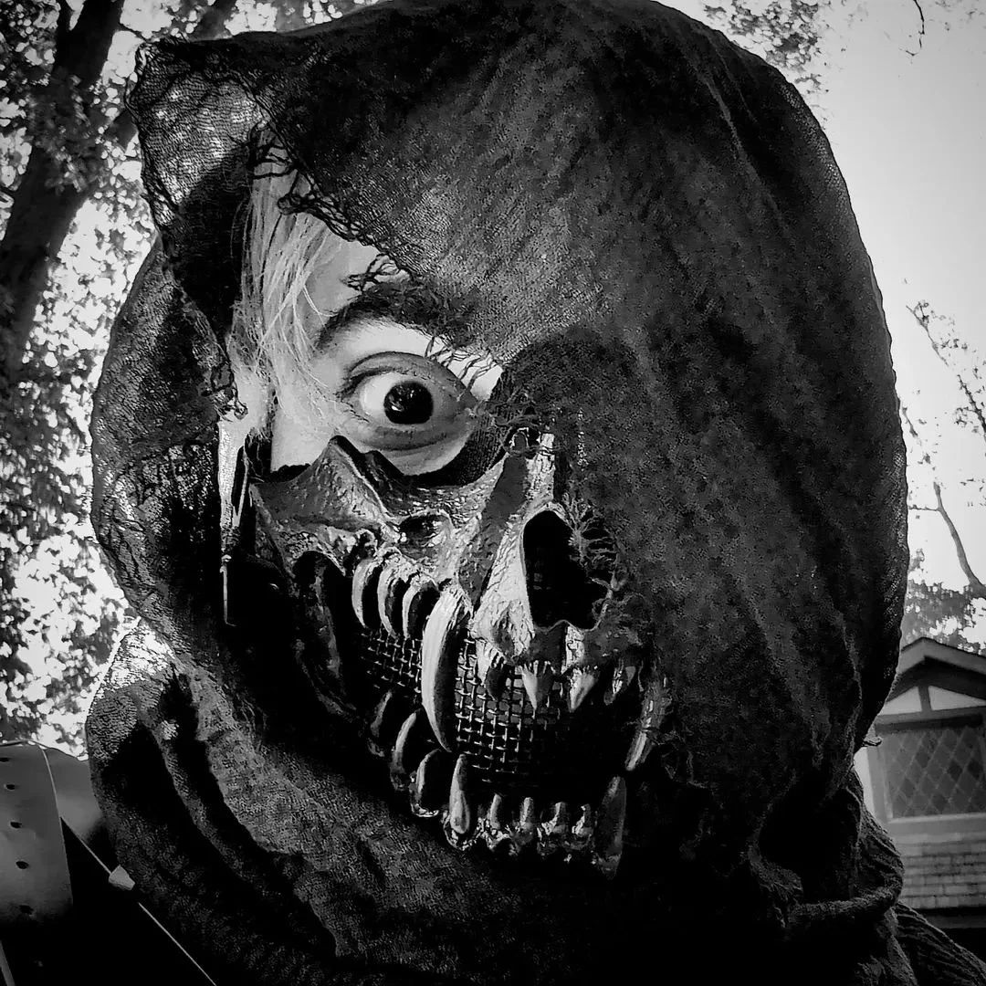 A person wearing a skull mask with an eye behind the mask, surrounded by a tattered black hood. The scene appears to be outdoors with trees and a building in the background, in black and white.