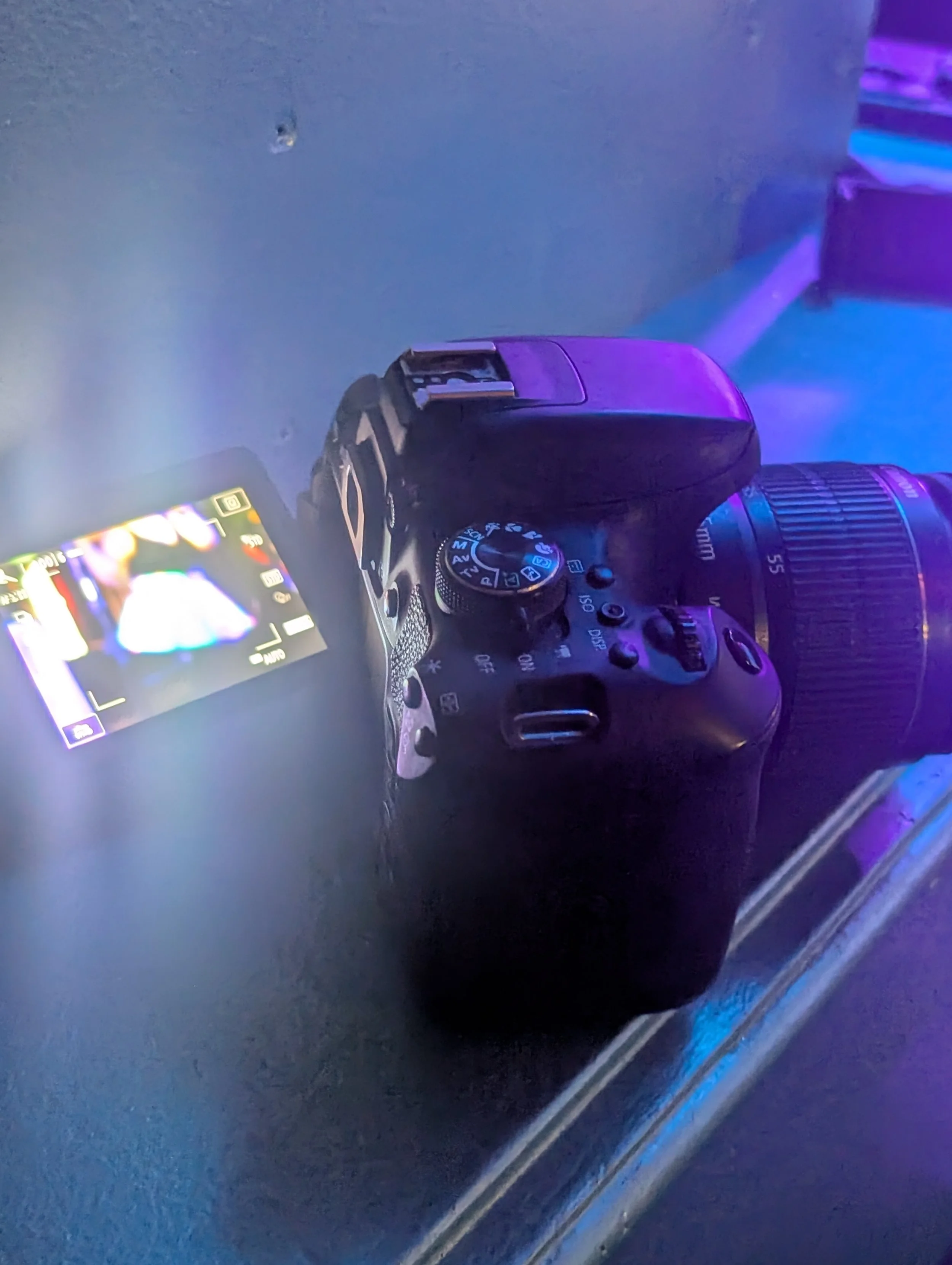 A digital camera with a lens attached, shown from a side angle on a dark surface, with colorful lighting reflecting off it.