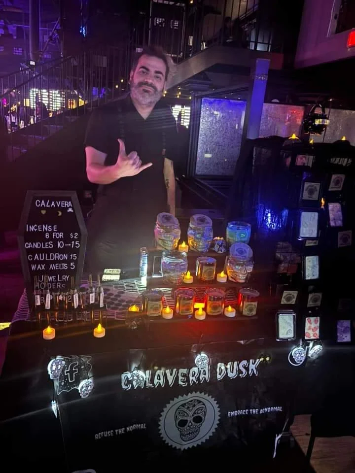 A person behind a small table with candles, soaps, and incense at a nighttime event. The table has a sign reading 'Calavera Dusk' and offers items like candles, incense, and wax melts. The person is making a shaka hand gesture and smiling.