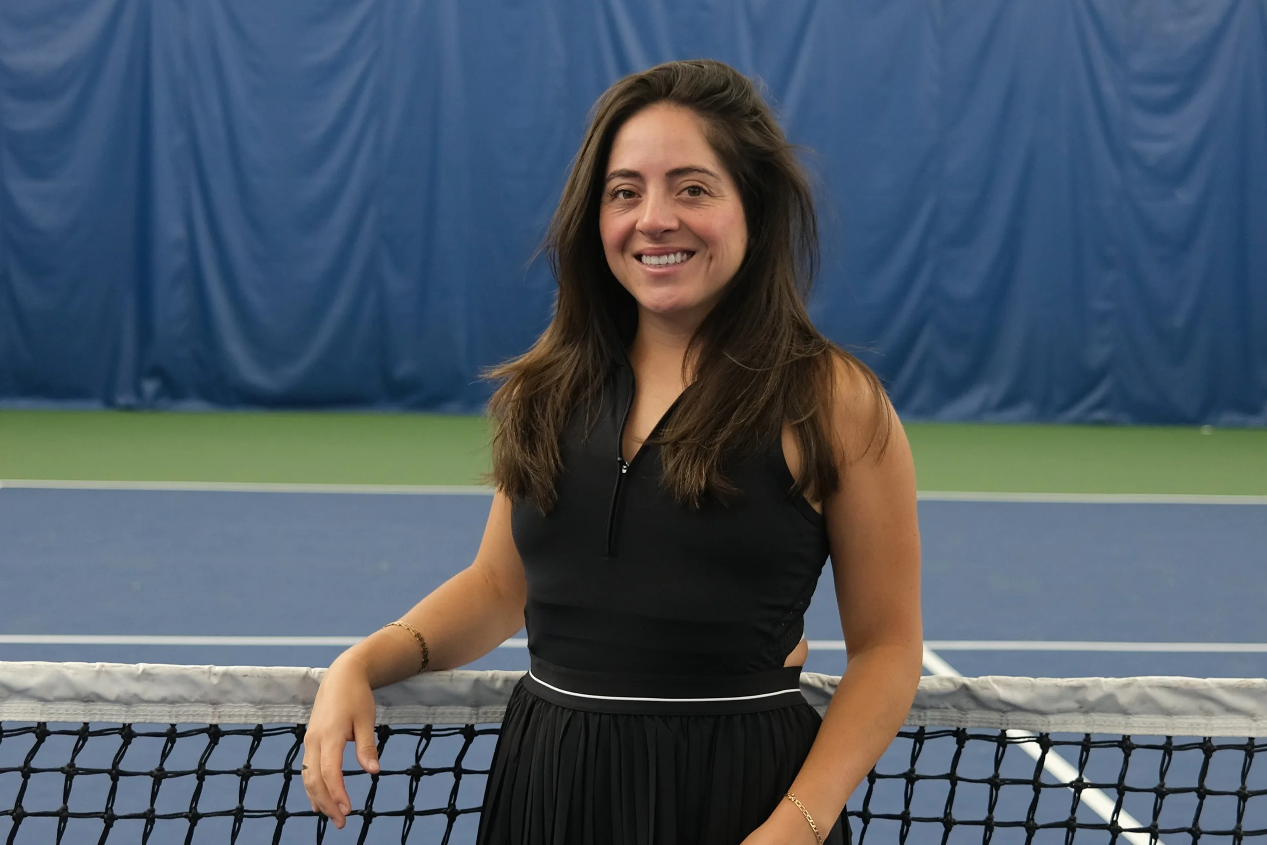 A woman with long brown hair standing on a tennis court, smiling at the camera, wearing a black sleeveless top and a skirt, with a tennis net in front of her and blue curtains in the background.