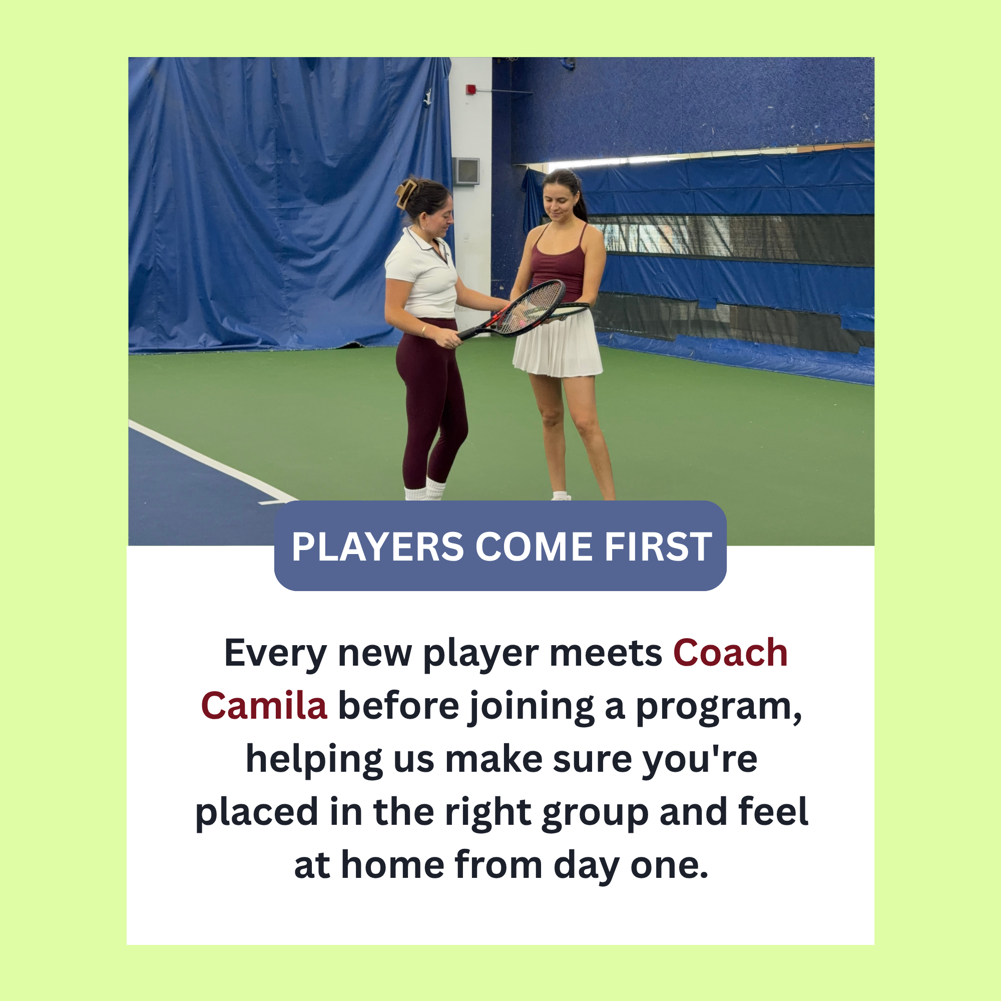 Two women stand on an indoor tennis court, one holding a tennis racket, while the other appears to be coaching or instructing her. They are wearing athletic clothing, and the court has blue walls with a large blue curtain in the background.