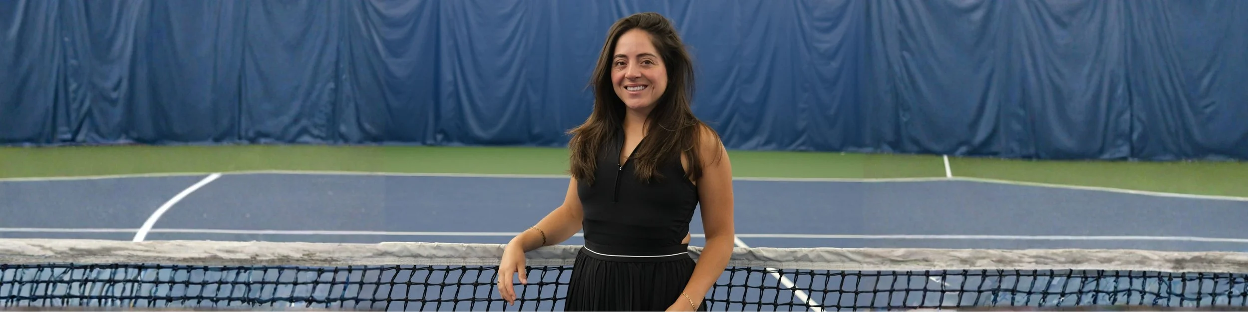 A woman standing in front of a tennis net on an indoor tennis court, smiling at the camera.