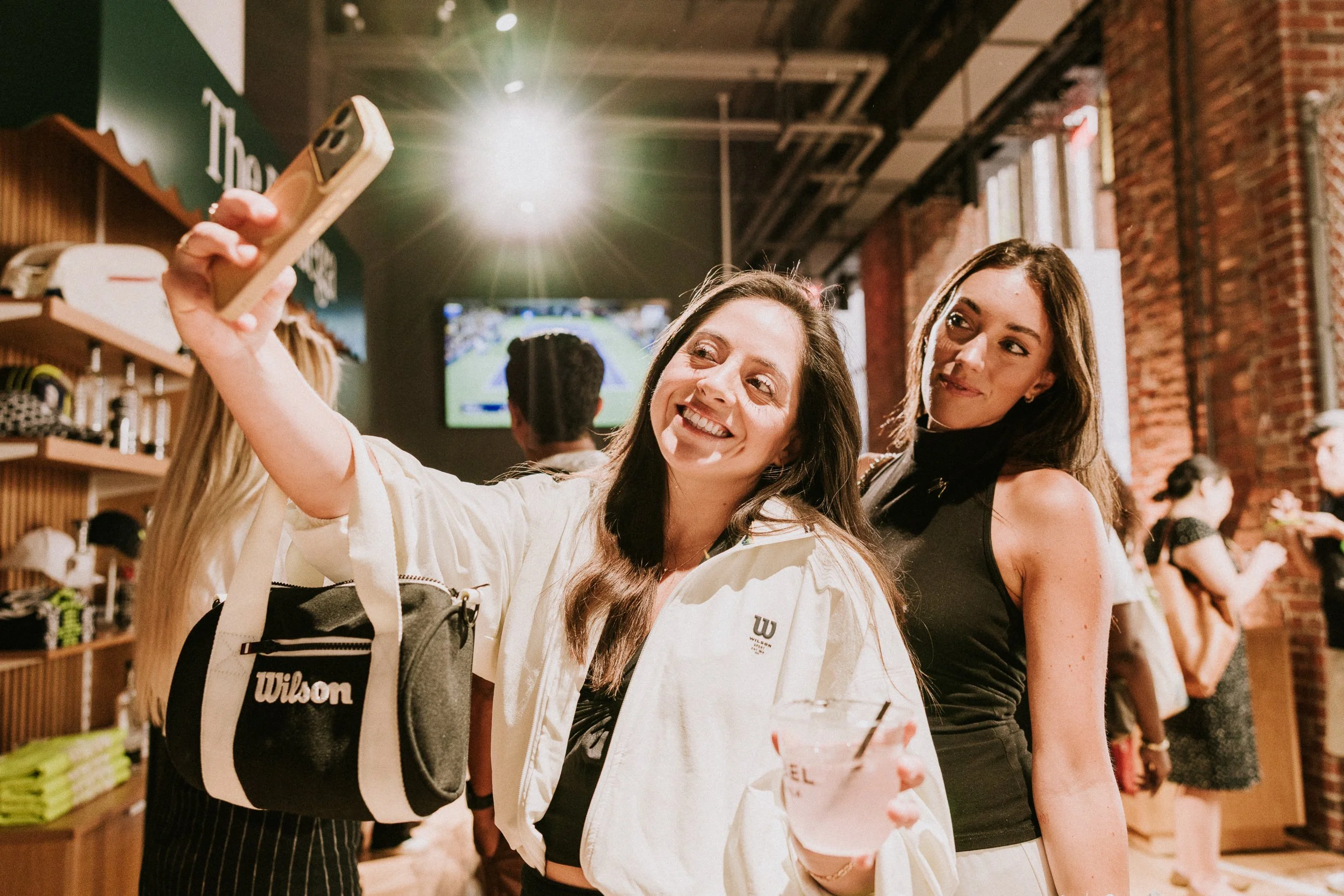 Two women smiling and taking a selfie inside a store that sells tennis equipment and apparel. One woman is holding a smartphone, and the other is looking at the camera. There are shelves with shoes and tennis gear in the background, and a television displaying a tennis match.