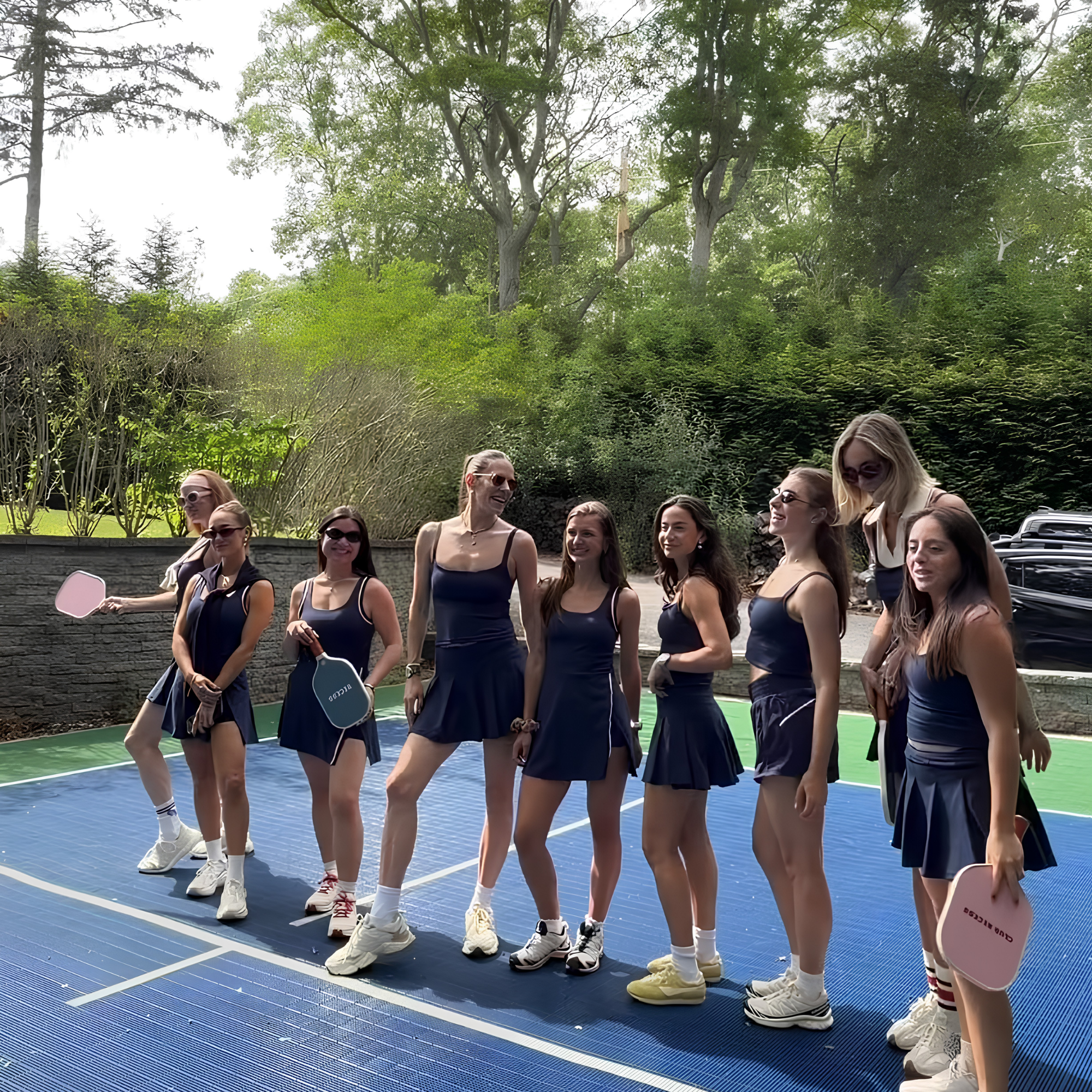 Women standing in a row on a blue basketball court outdoors, wearing matching navy blue outfits and sunglasses, with trees and parked cars in the background.