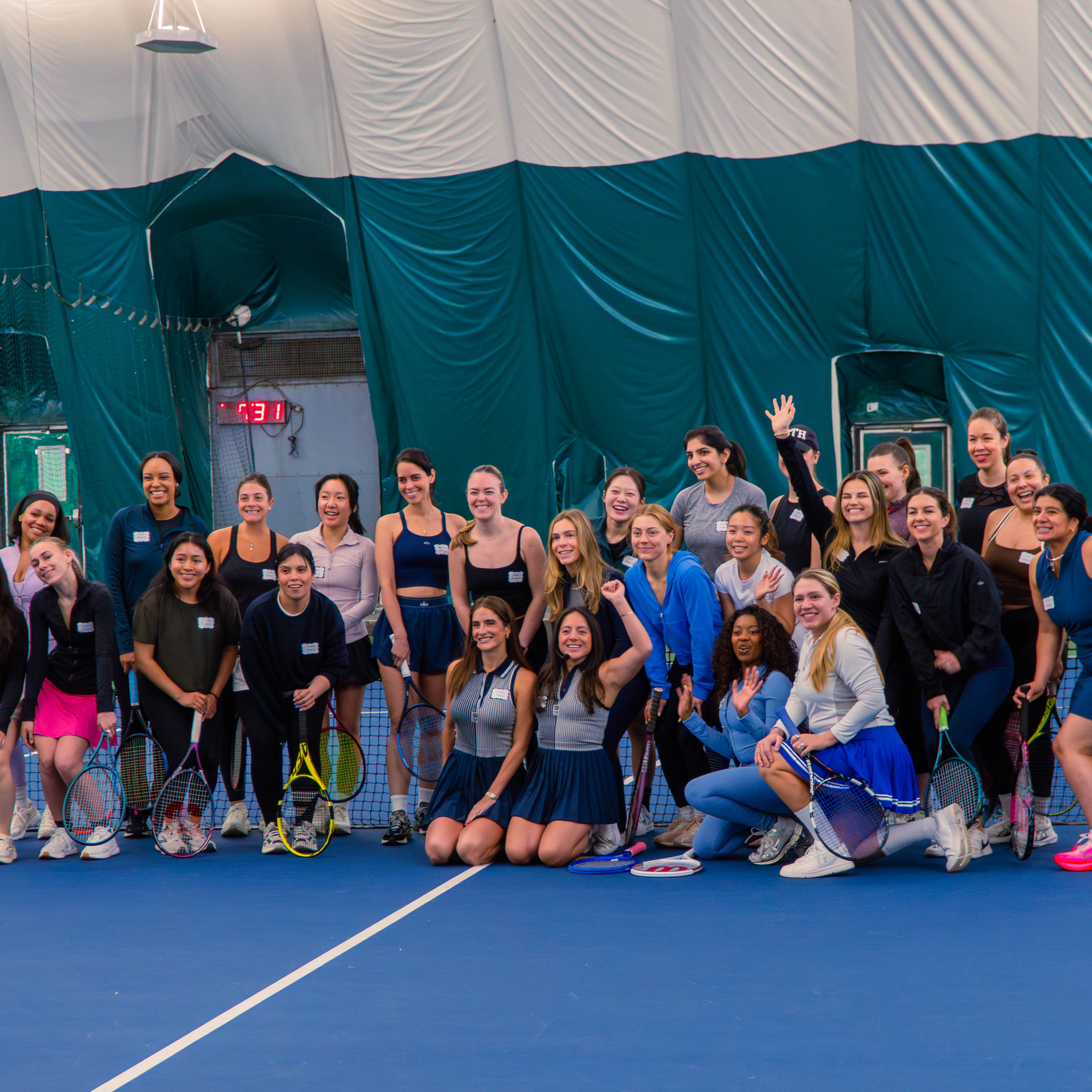 Group of young women, some holding tennis rackets, smiling and posing on a tennis court at an indoor sports facility with a blue floor and white lines, and a large draped tent-like ceiling.