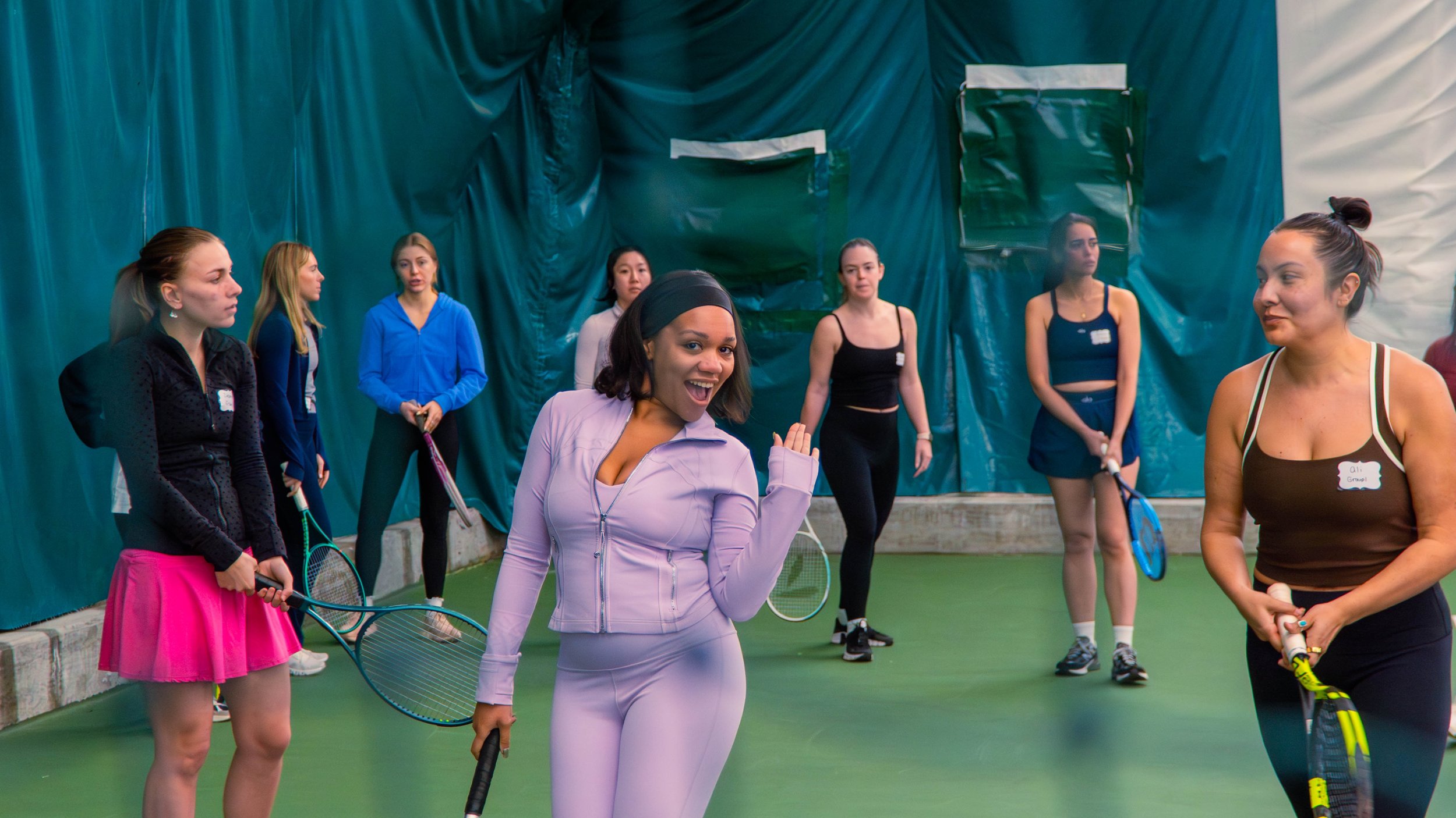 Group of women on an indoor tennis court, some holding tennis rackets, with a woman in lavender sportswear smiling and posing in the foreground.