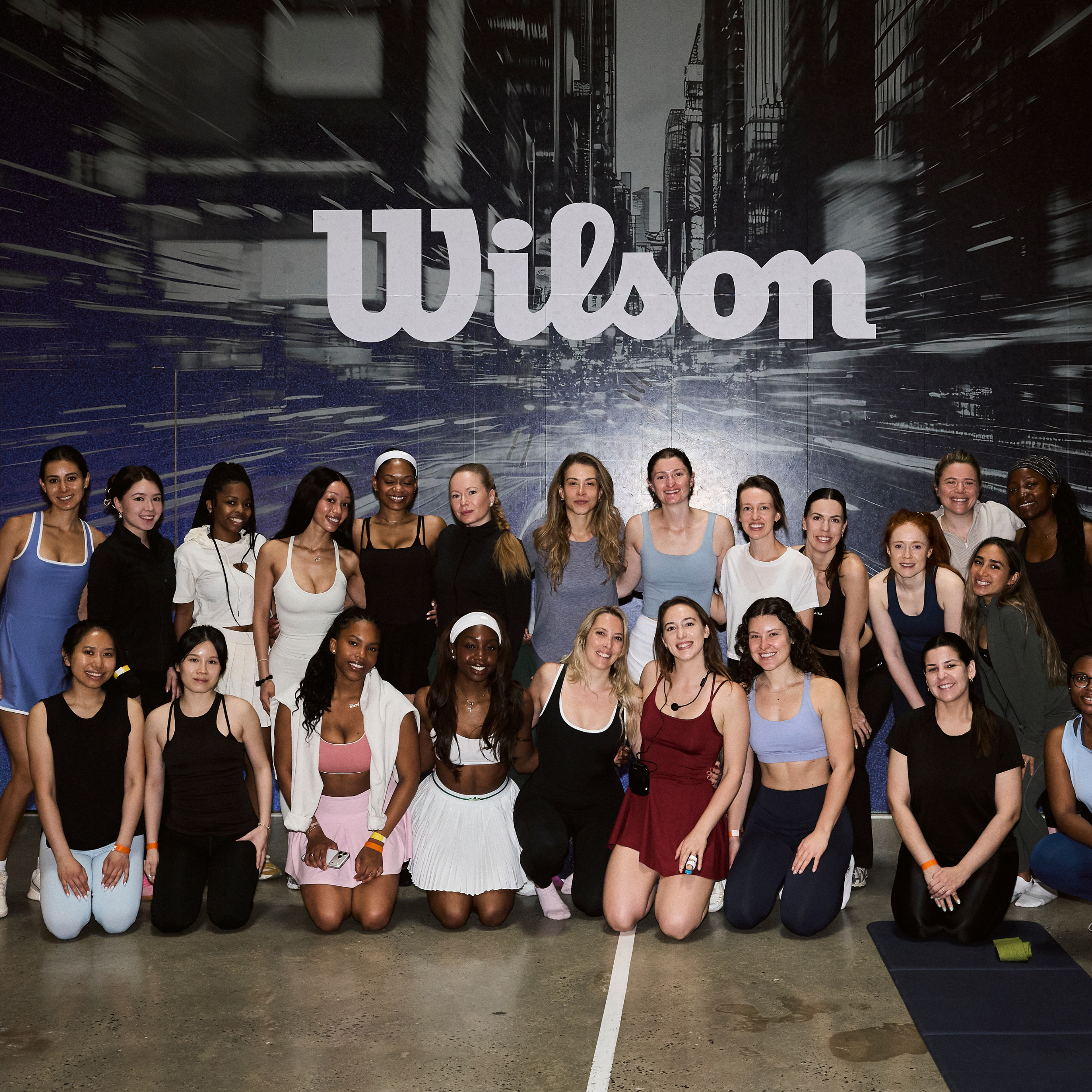 Group of young women smiling and posing together in athletic wear on an indoor sports court with a large sign that says 11:00 am in the background.
