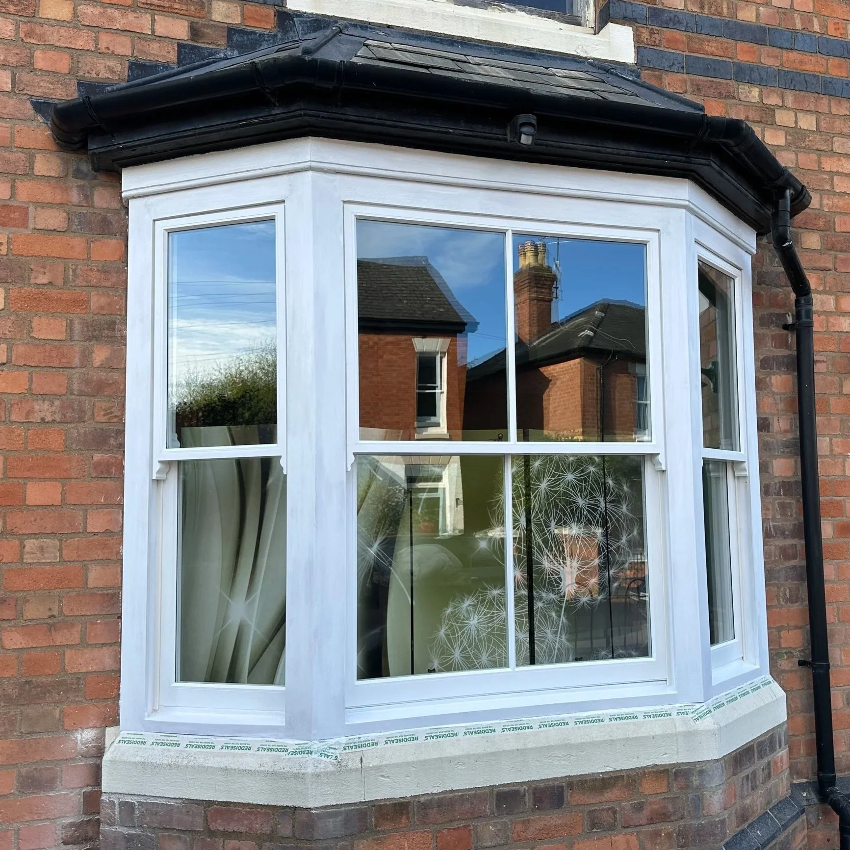 A new white bay window installed on a brick house, with protective tape at the bottom reading 'REDFIELD'S'. The window has four sections with clear glass, reflecting neighboring houses and the sky.