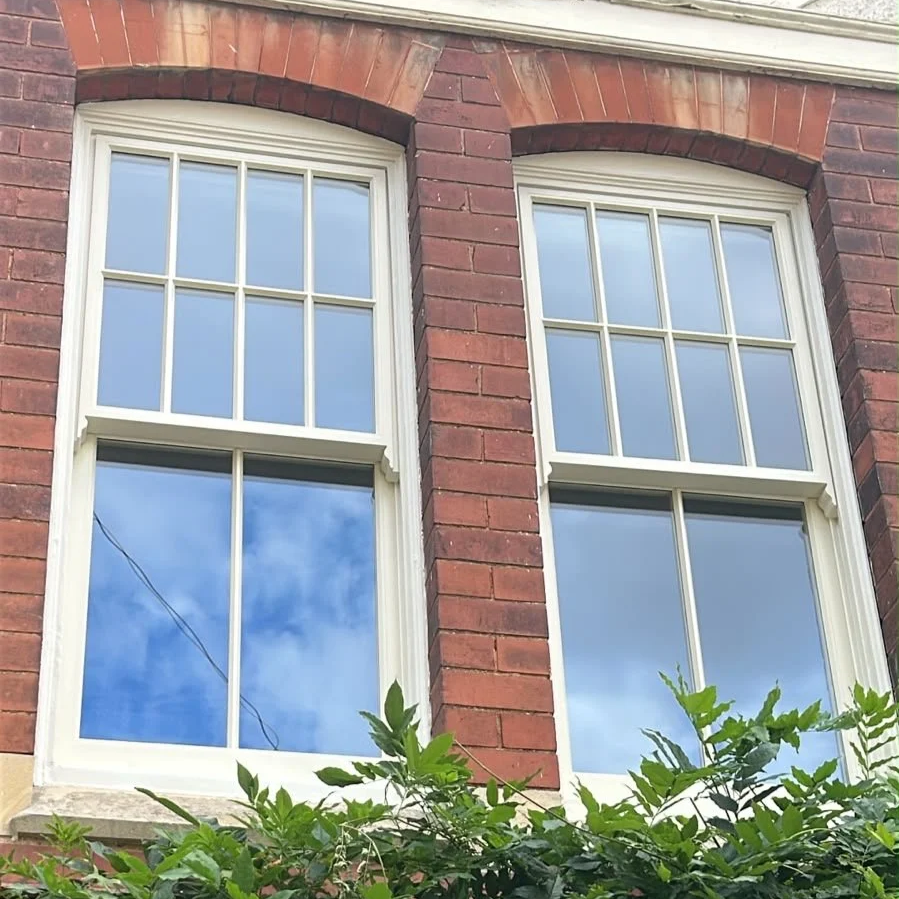 Two large double-hung windows with white wooden trim and decorative stained brick arches in a red brick building, with greenery at the bottom and reflections of a blue sky and clouds in the glass.