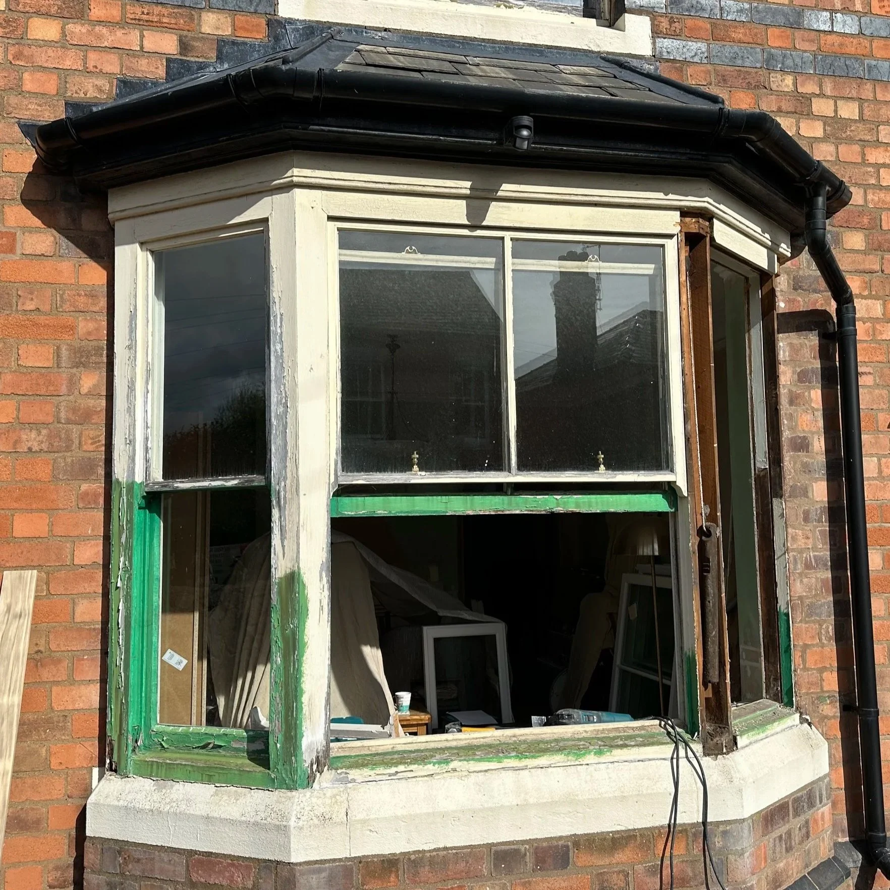 A brick house exterior with a bay window undergoing renovation; the window frame is partially removed, revealing the interior walls and window sills, with tools and construction materials inside.