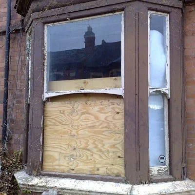 A brown wooden bay window boarded up with plywood, with a small section of window still visible on the right side, and a reflective glass window showing the sky and a building across the street.