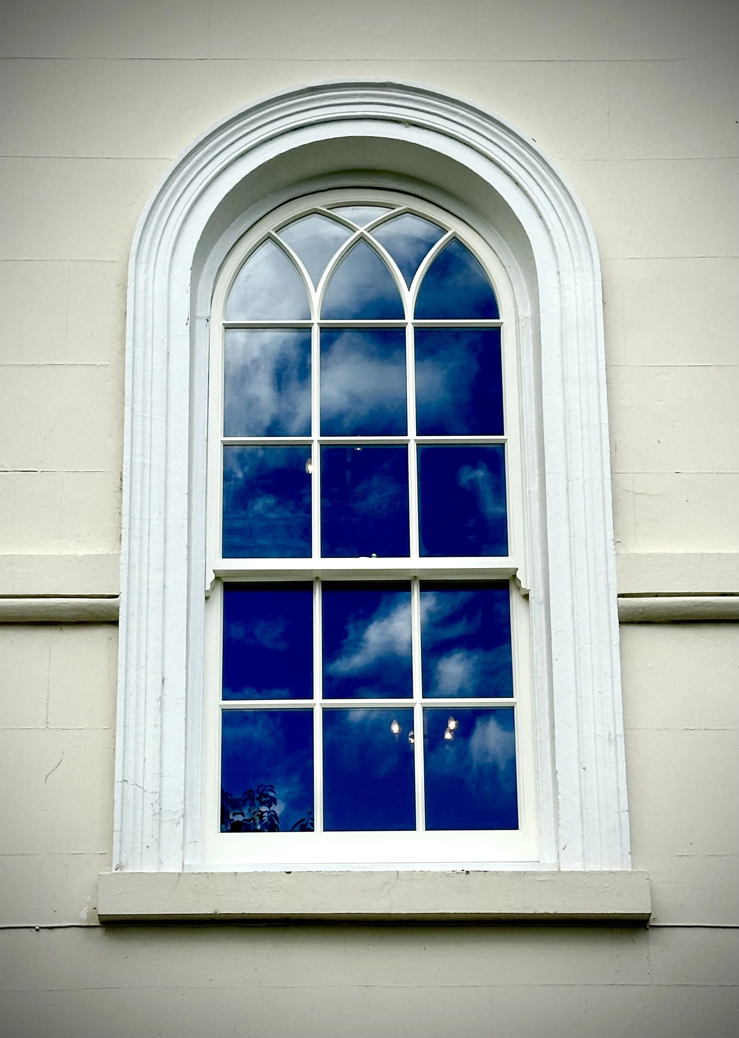 A large arched window with multiple panes, reflected blue sky with clouds, on a beige building exterior.