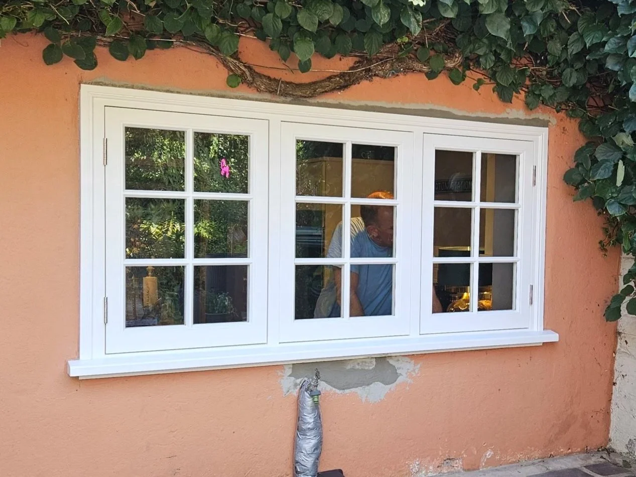 A white-framed window with three sections installed on a peach-colored wall, with some repair work visible beneath it and climbing green vines above.