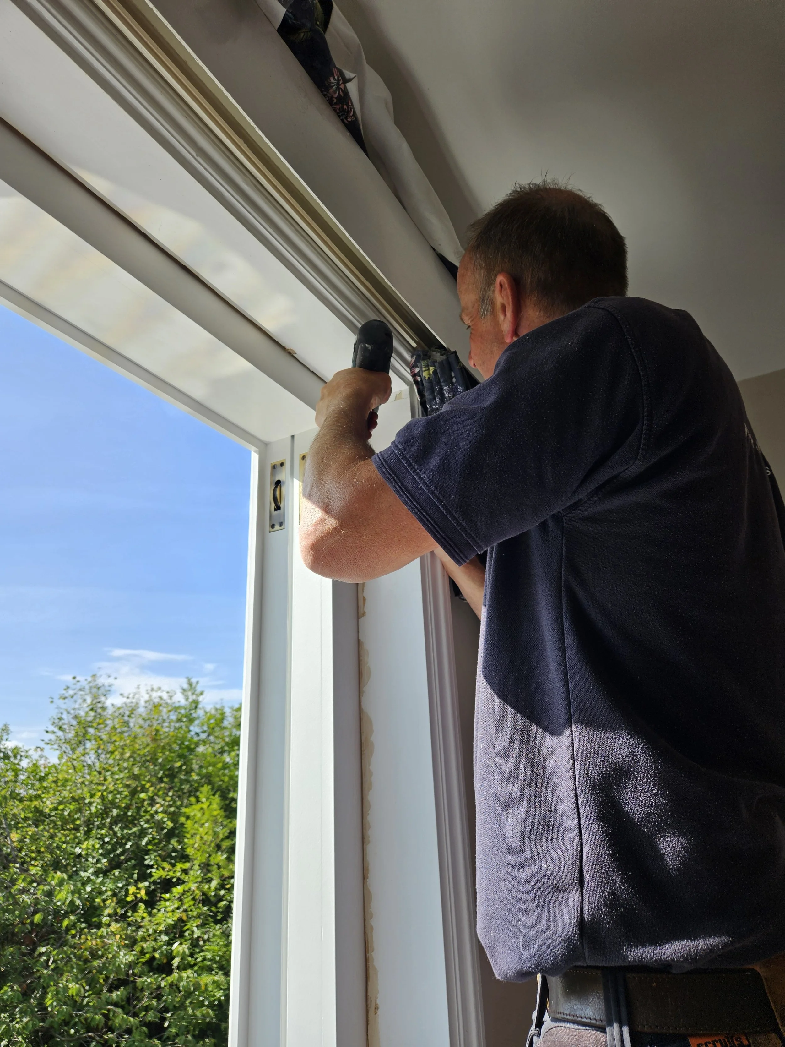 A man working on a window frame with a screwdriver during daylight, with green trees and a blue sky outside.