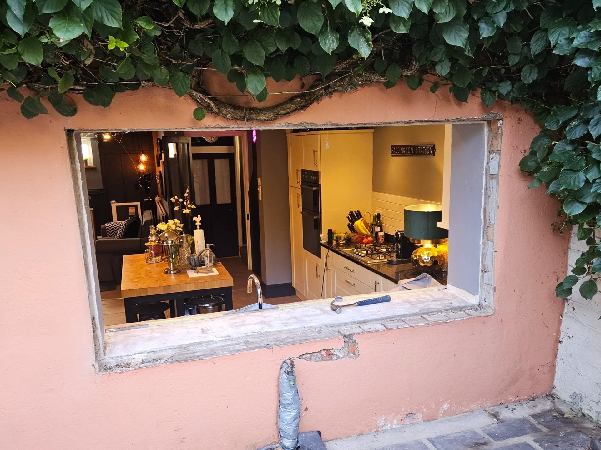 Interior view of a kitchen with renovation in progress, seen through an unfinished window opening in a pink exterior wall. The kitchen has yellow cabinets, a black stove, a countertop with various items, and warm lighting.