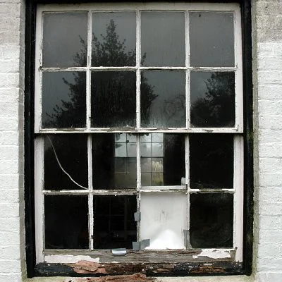 Old, broken window with multiple panes, some missing or cracked, on a brick building. Reflection of trees and sky visible in the glass.