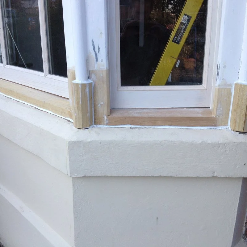 Close-up of a window and door frame under construction, with wooden trim and unfinished white wall.