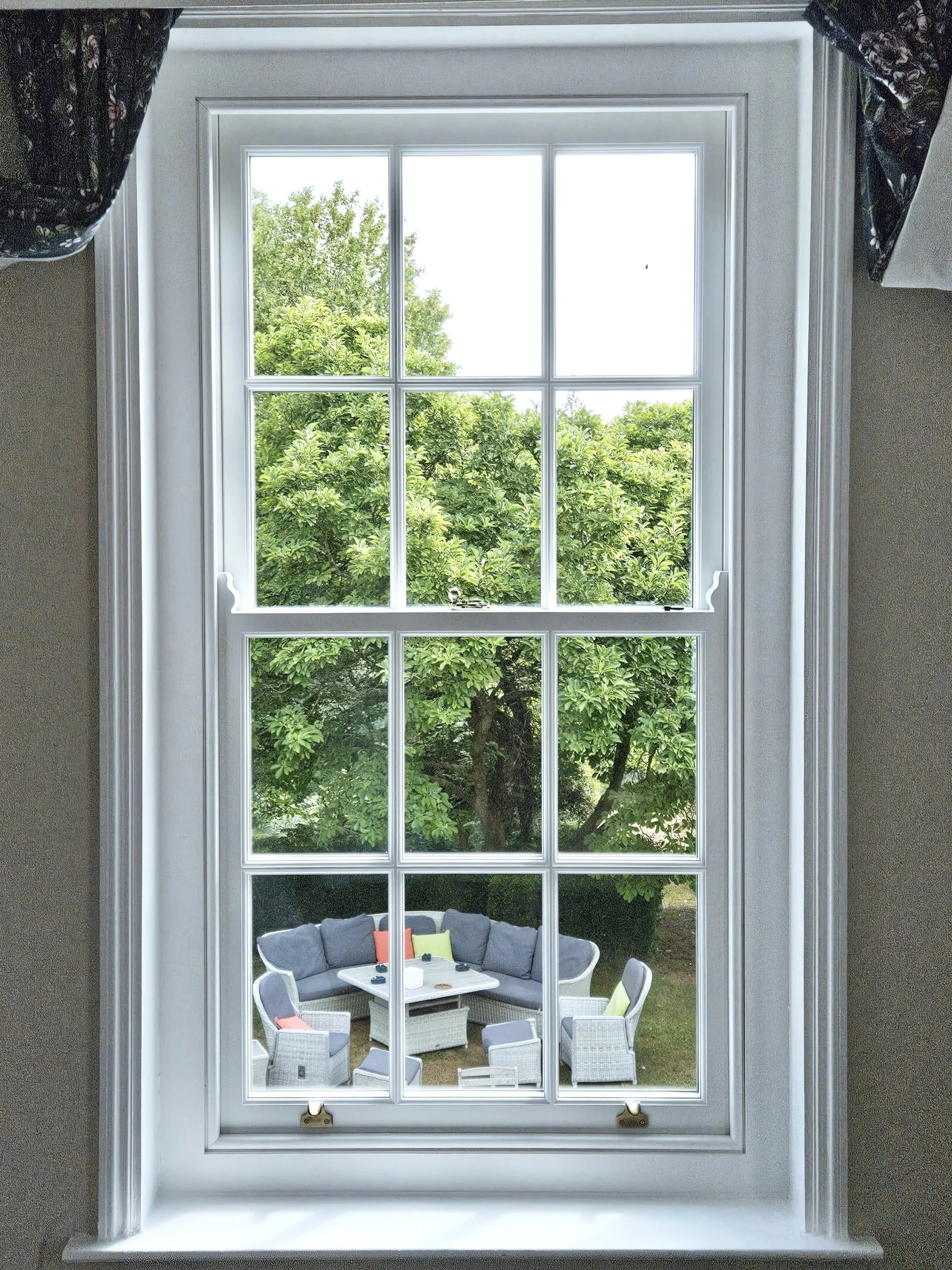 Looking through a large window with nine panes, displaying a backyard patio with a gray outdoor sofa, chairs, and a small table surrounded by lush green trees.