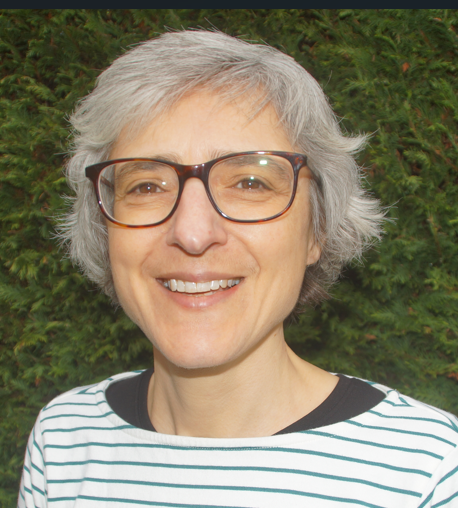 Smiling woman with short gray hair and glasses, wearing a black and white striped shirt, standing outdoors in front of green foliage.