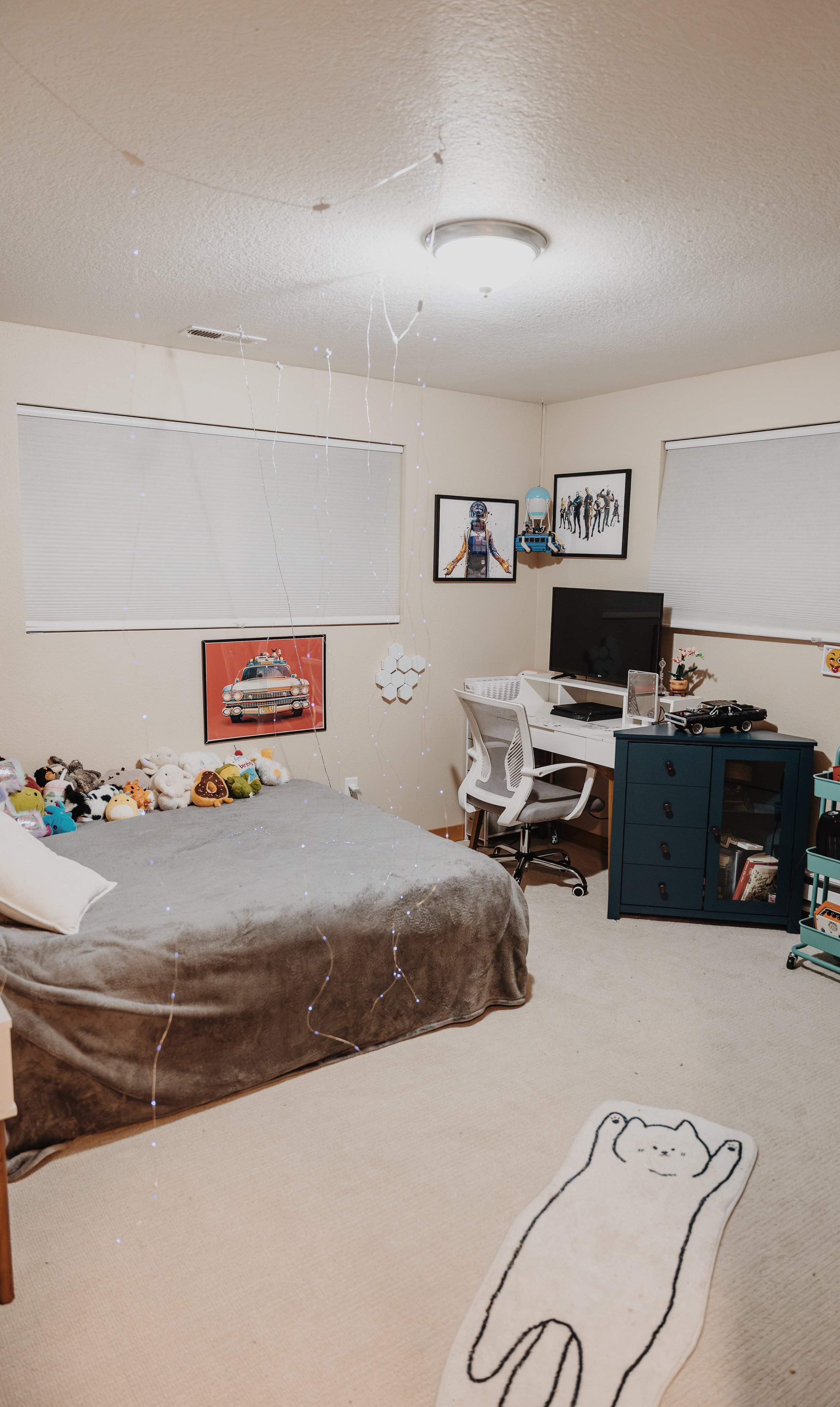 A bedroom with a bed covered in plush toys, a white desk with a computer and TV, framed wall art, a blue storage cabinet, a black and white cat rug on beige carpet, and string lights hanging from the ceiling.