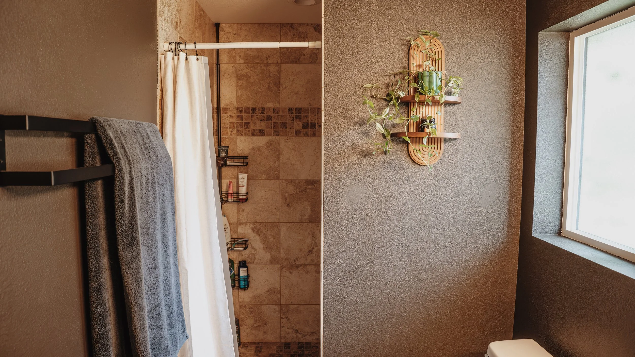 A bathroom with a window on the right wall, a gray towel hanging on a rack, a shower area with beige tile and a white curtain, and a wall-mounted wooden shelf with potted plants.