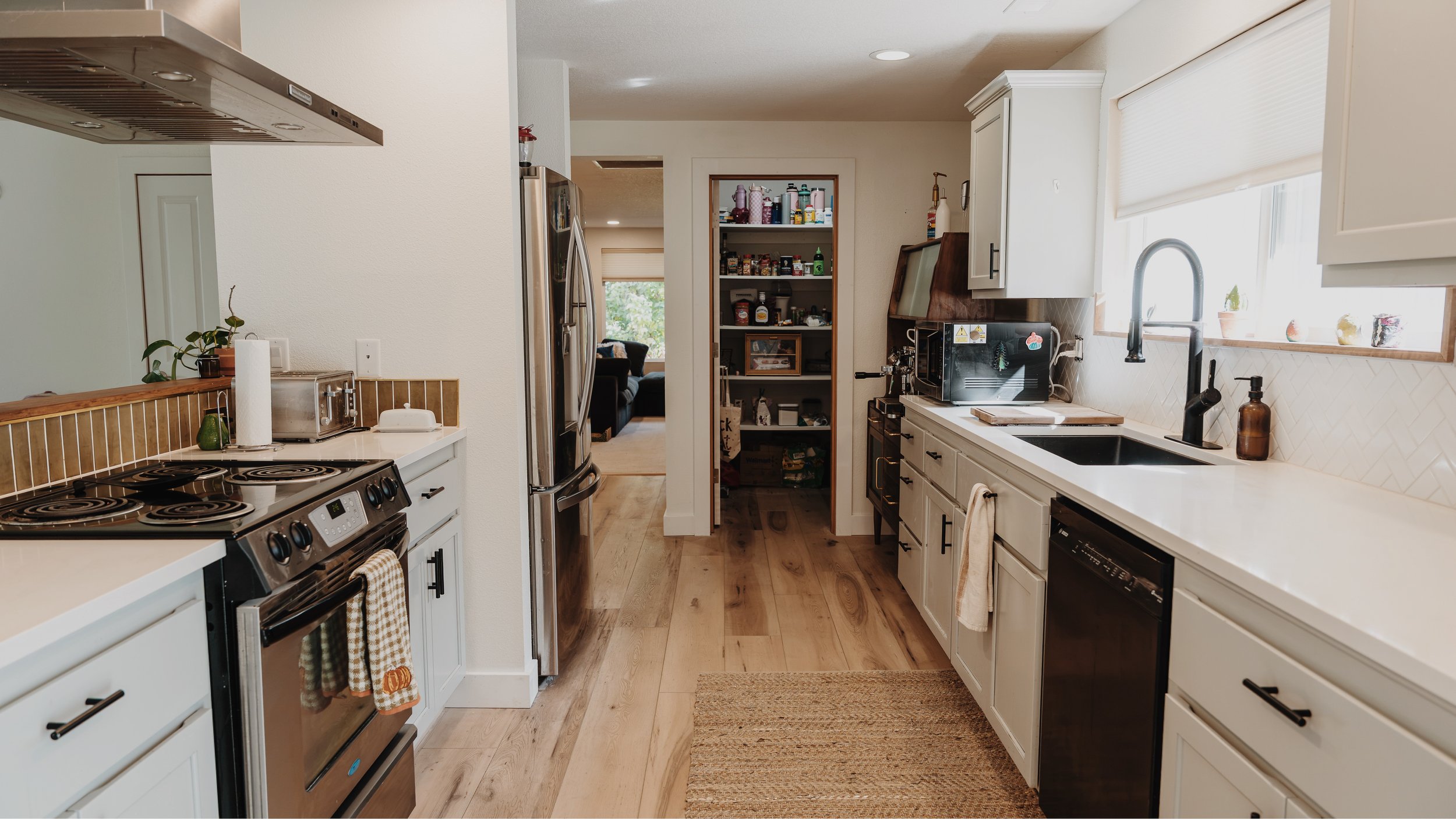 Kitchen with white cabinets, black sink, and stainless steel appliances. There is a window with a white blind, a countertop with a toaster and potted plants, and a pantry with various food containers. A living room with a sofa is visible through a doorway.