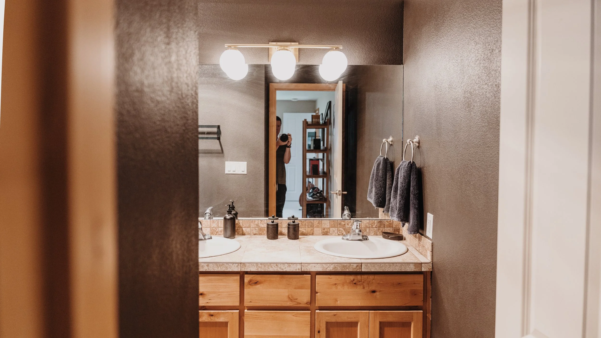 Bathroom mirror reflecting a person taking a photo, with a double sink vanity, dark gray towels on the wall, and wooden cabinets below the counter
