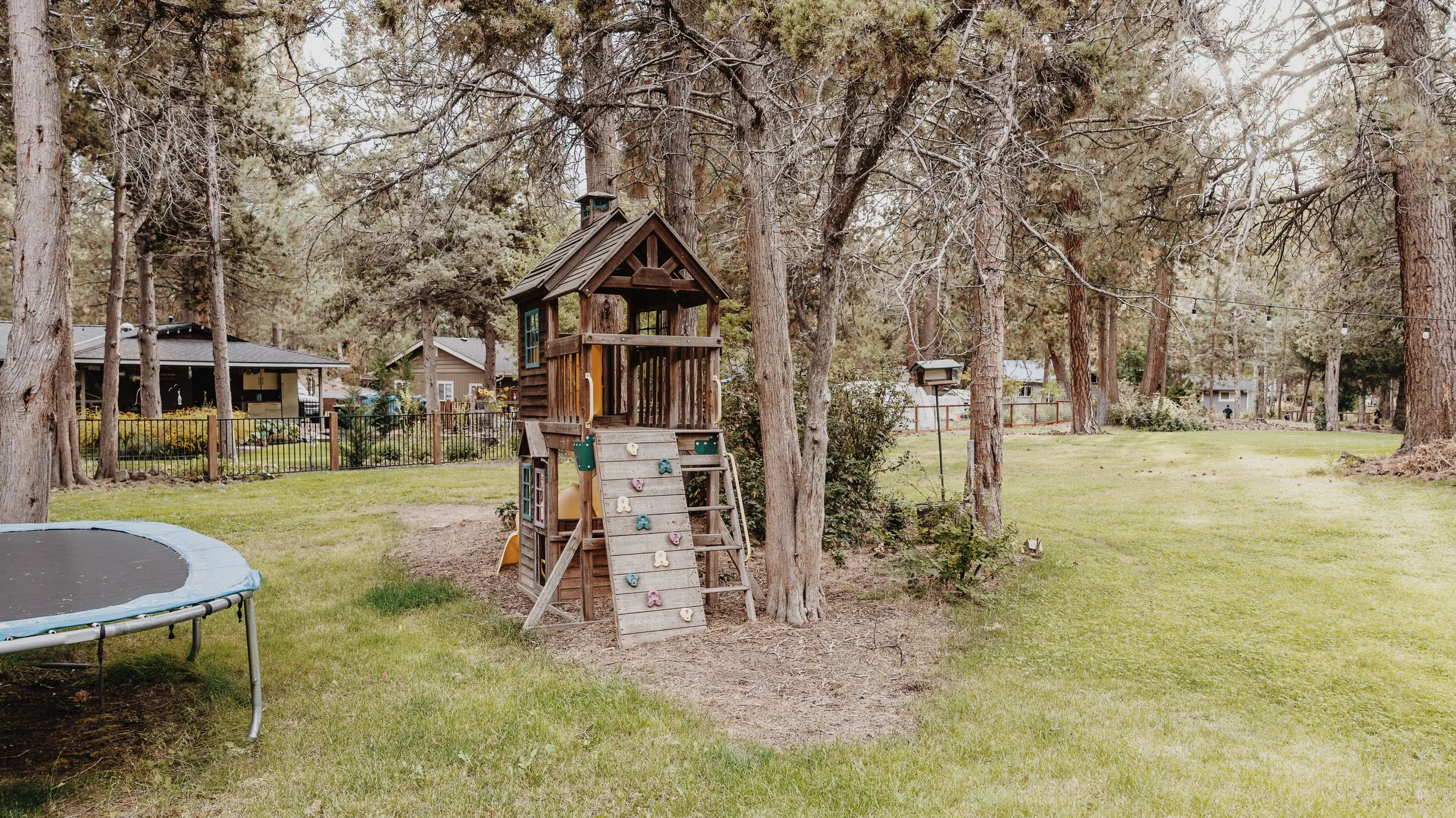 A backyard with a wooden playhouse, a trampoline, and large trees underscored by a grassy lawn, residential houses, and string lights.