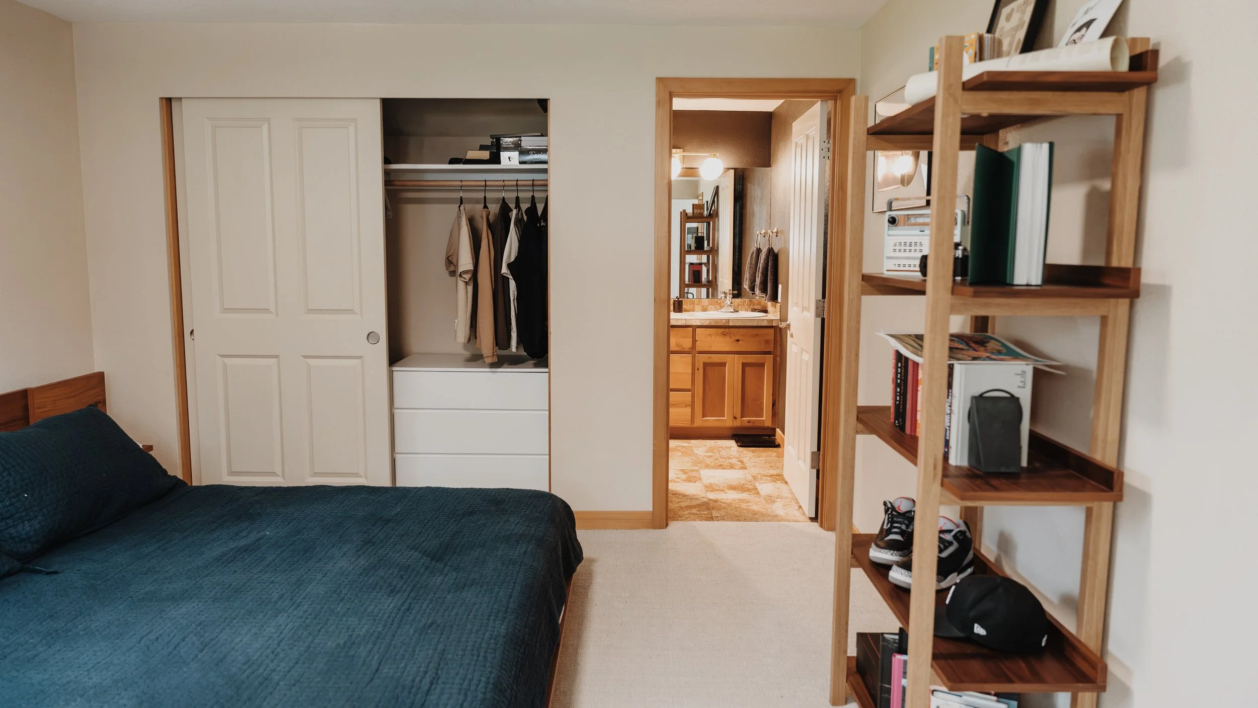 View of a bedroom with a closet, doorway to a bathroom, and a wooden bookshelf. The bed has a dark blue Cover. The closet has a few hanging clothes and drawers. The bathroom has wooden cabinetry and a mirror.