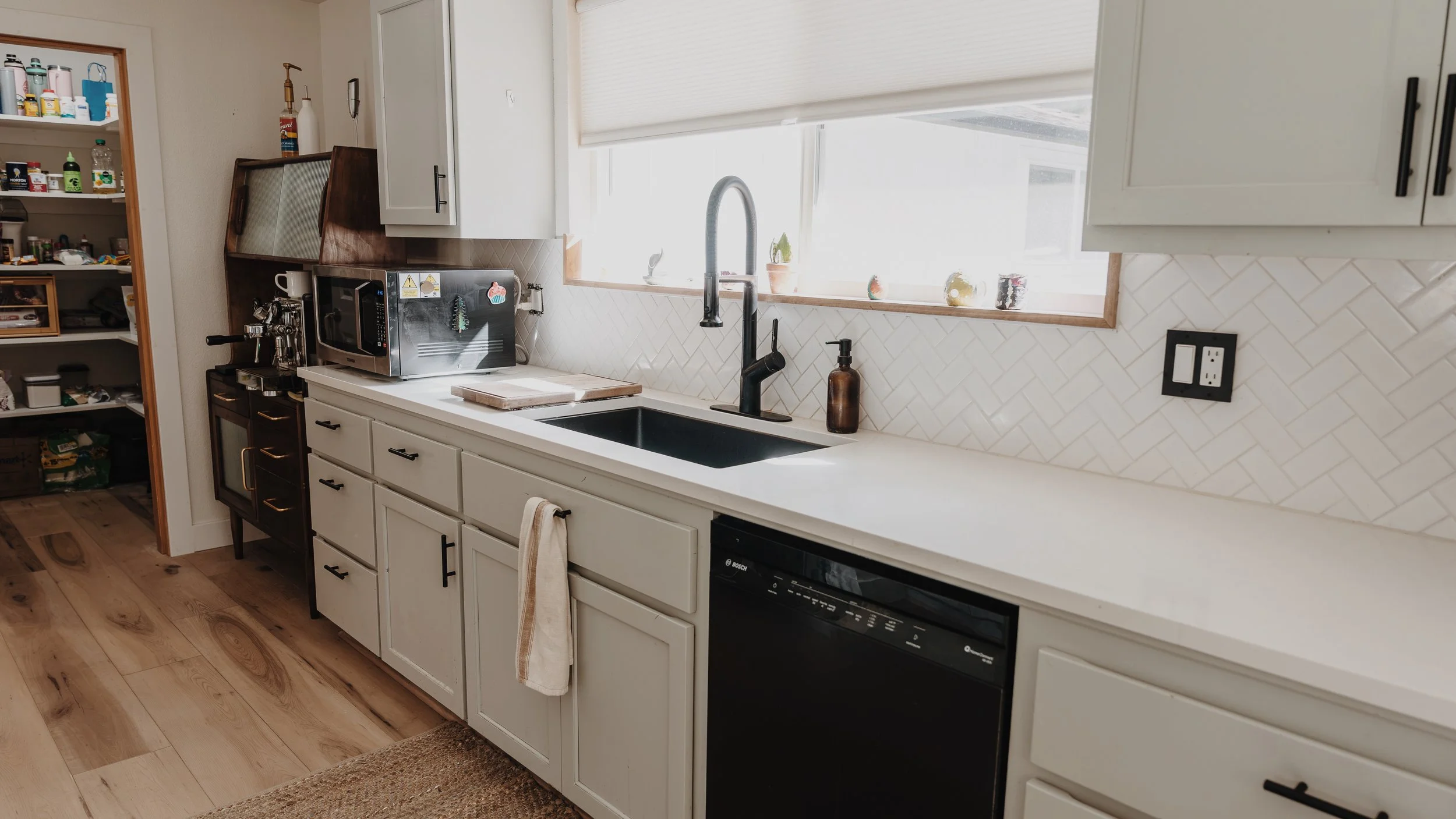 A kitchen with white cabinets, a black sink, a black dishwasher, and a white countertop. There is a window above the sink with a few small decorative plants. On the countertop, there are a brown soap dispenser, a microwave, and a cutting board. To the left, a shelf with various pantry items and a small black oven with a coffee machine on top are visible.