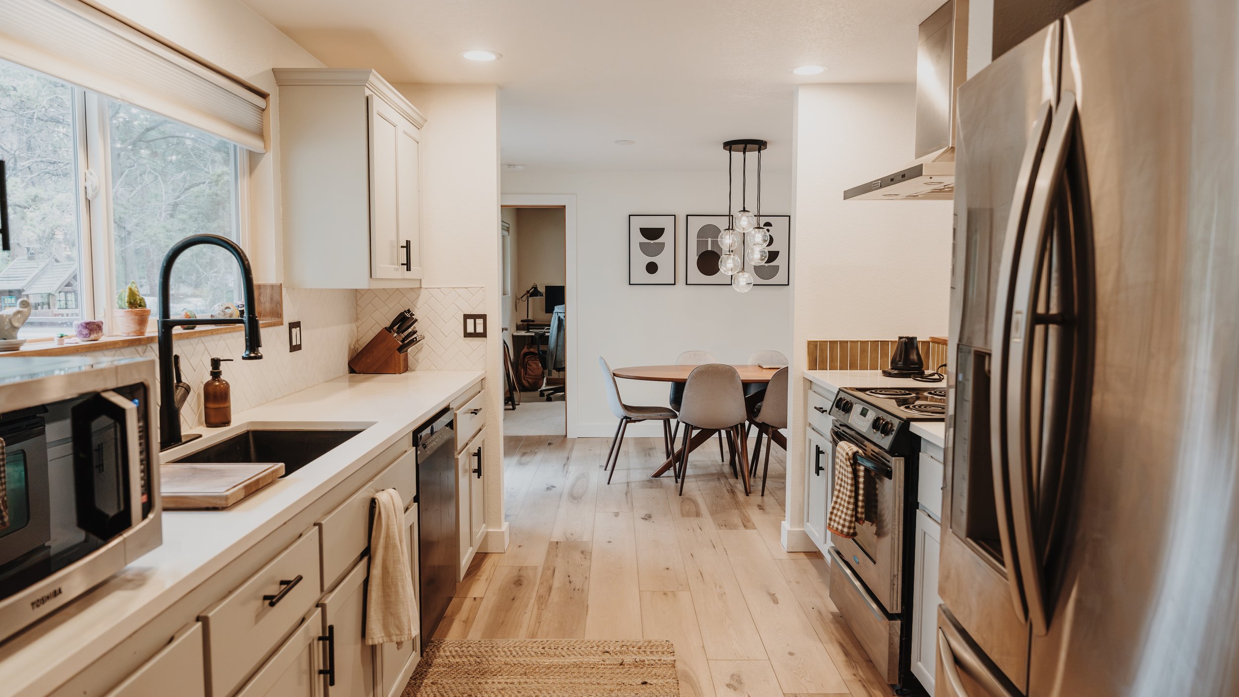 A kitchen with cabinets, a window overlooking trees, and a dining area with a round table and four gray chairs. The kitchen has stainless steel appliances, a black sink, and decorative wall art. There's a hanging light fixture with glass globes above the dining table.