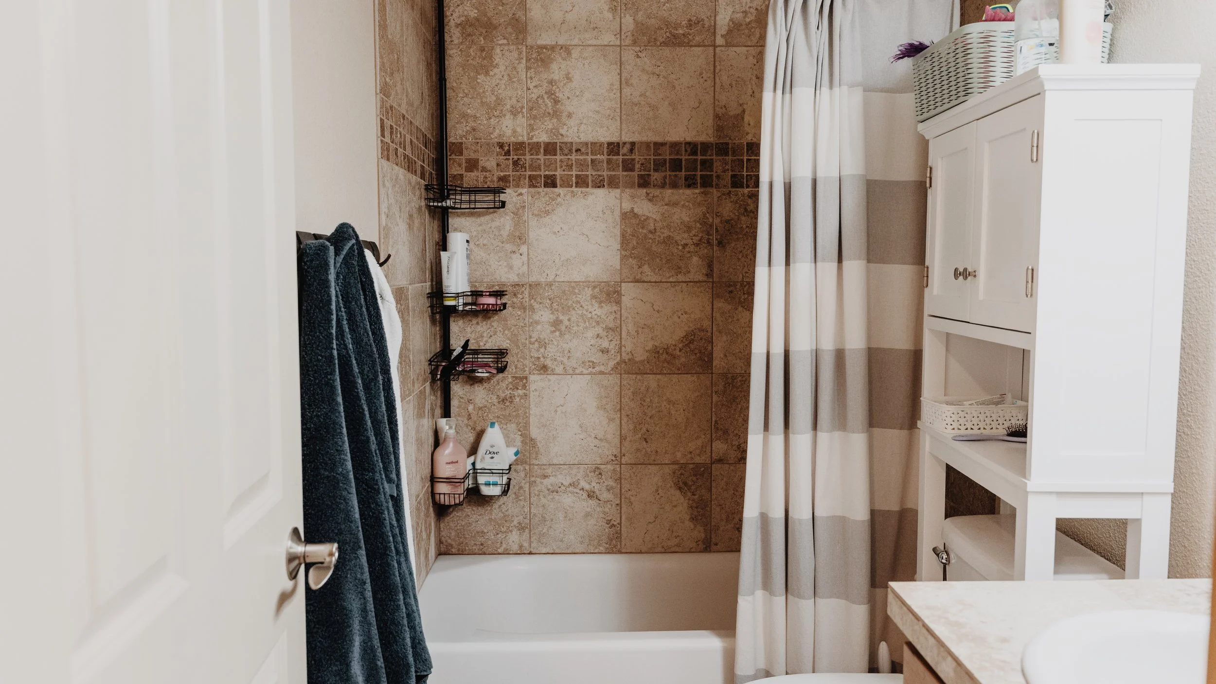 Bathroom with tiled shower area, shower curtain, and white bathroom cabinet with soap and toiletries.