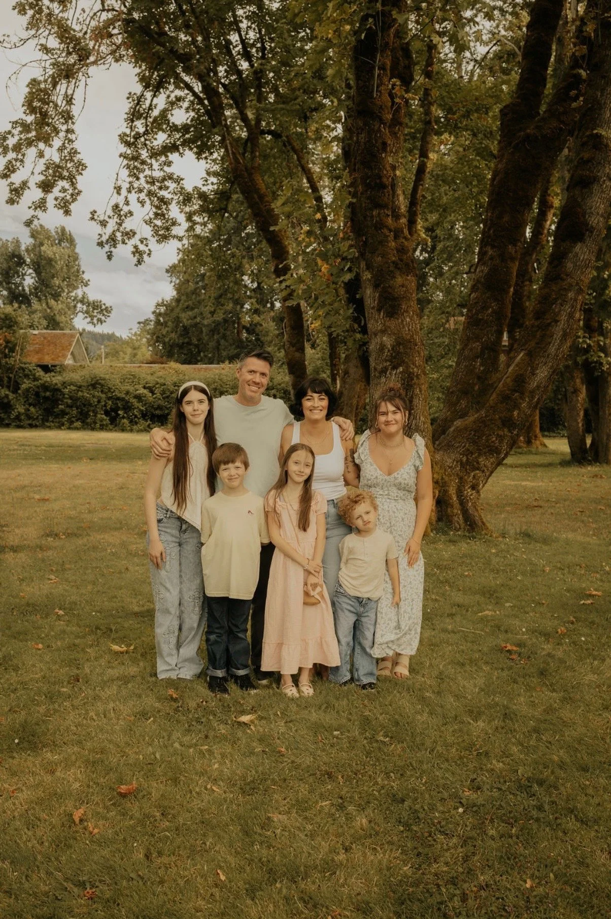 A family of seven standing in a park with large trees in the background. They are smiling and posing for the photo.